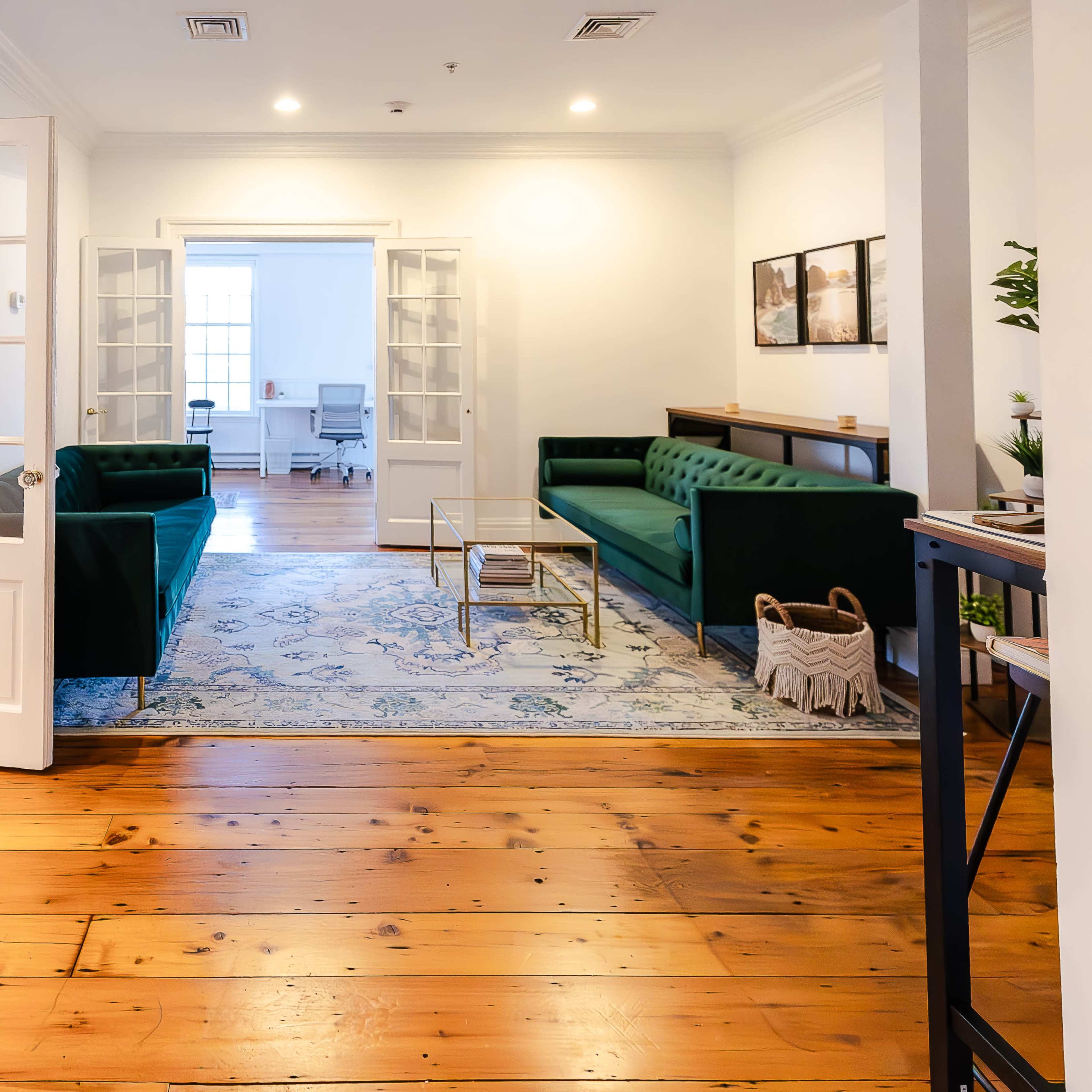 A well-lit office space featuring green sofas, a coffee table, and wooden flooring, with an adjacent room visible through open double doors.