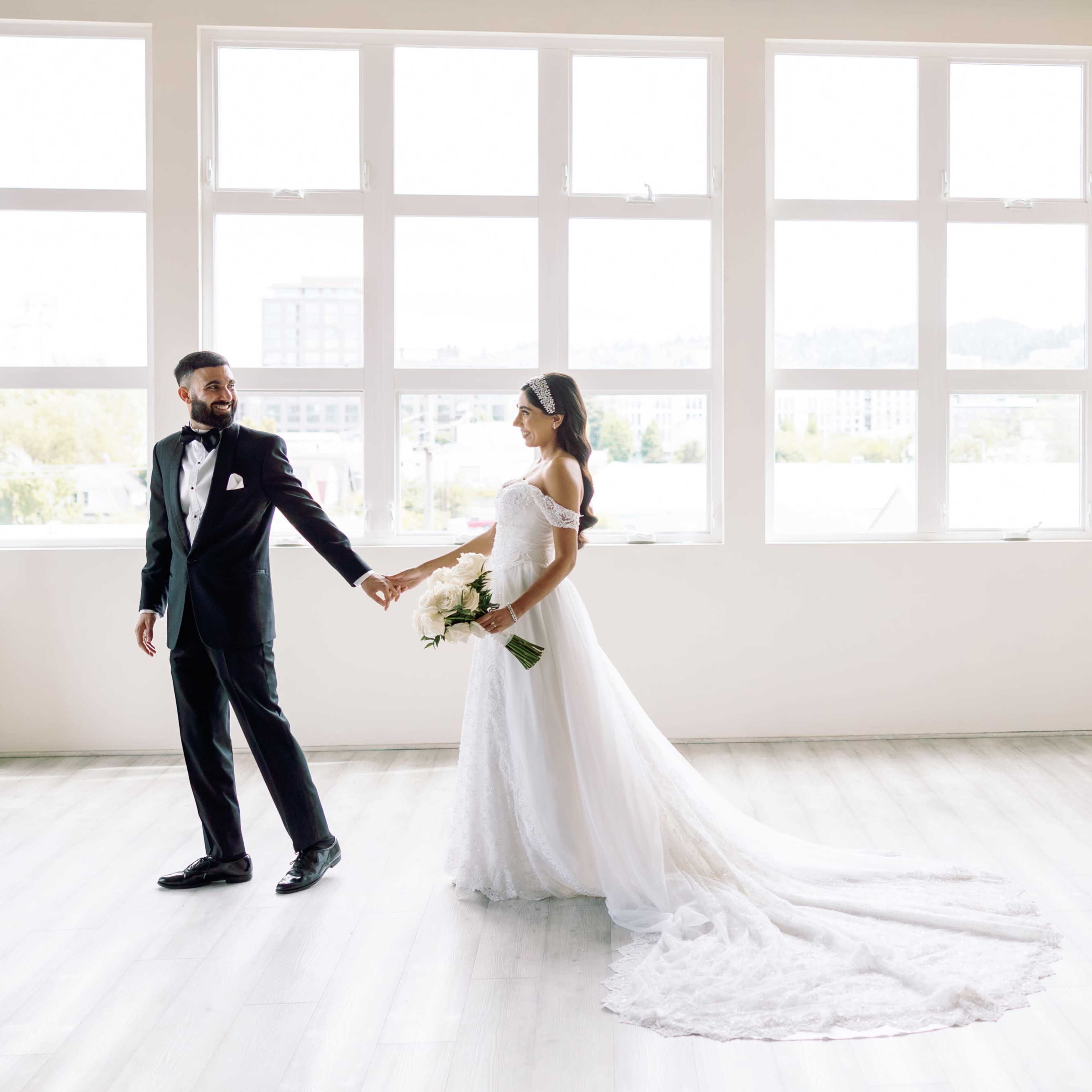 A bride and groom hold hands and walk together in a spacious, well-lit room with large windows.
