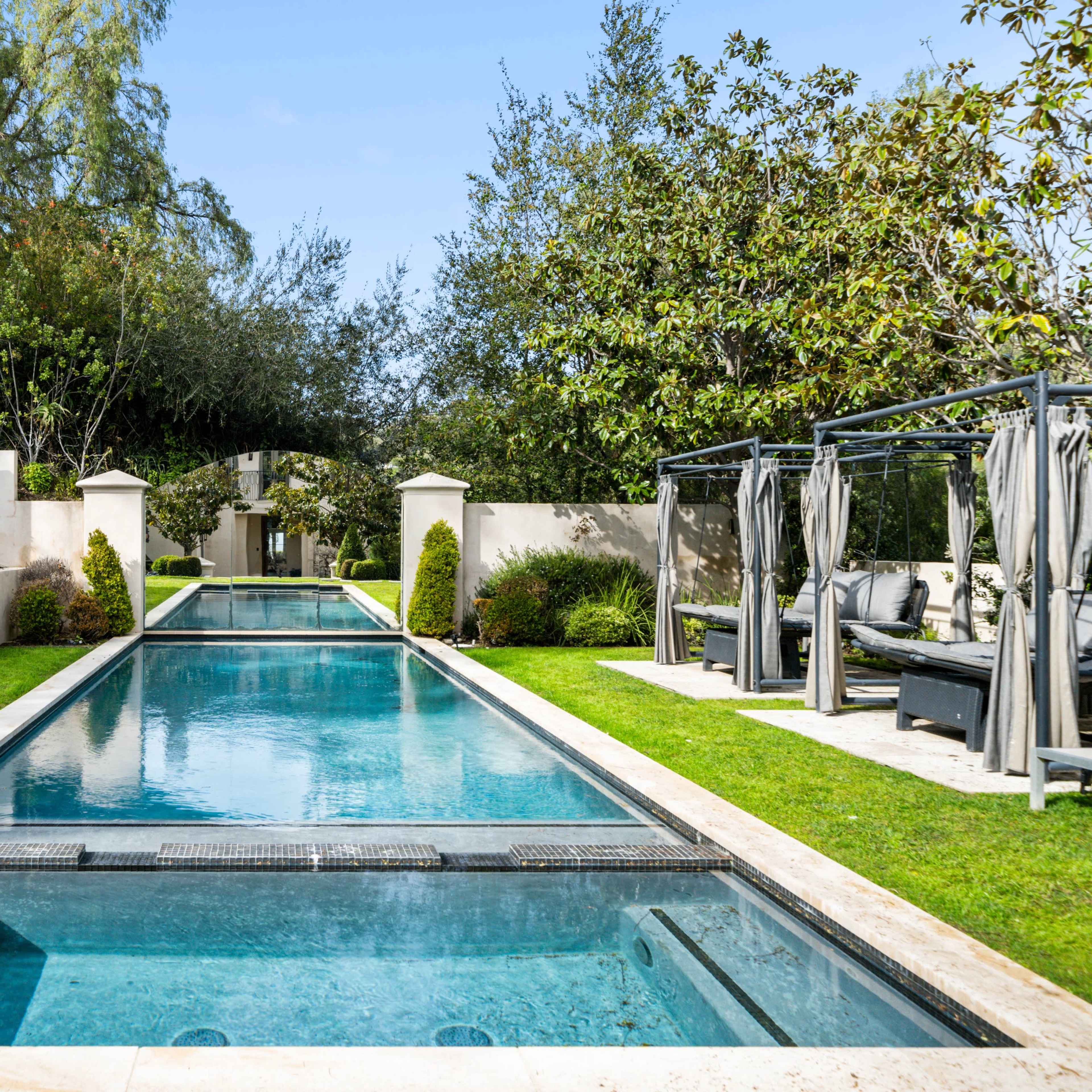 A landscaped outdoor area featuring a long, rectangular swimming pool flanked by greenery and lounge cabanas.