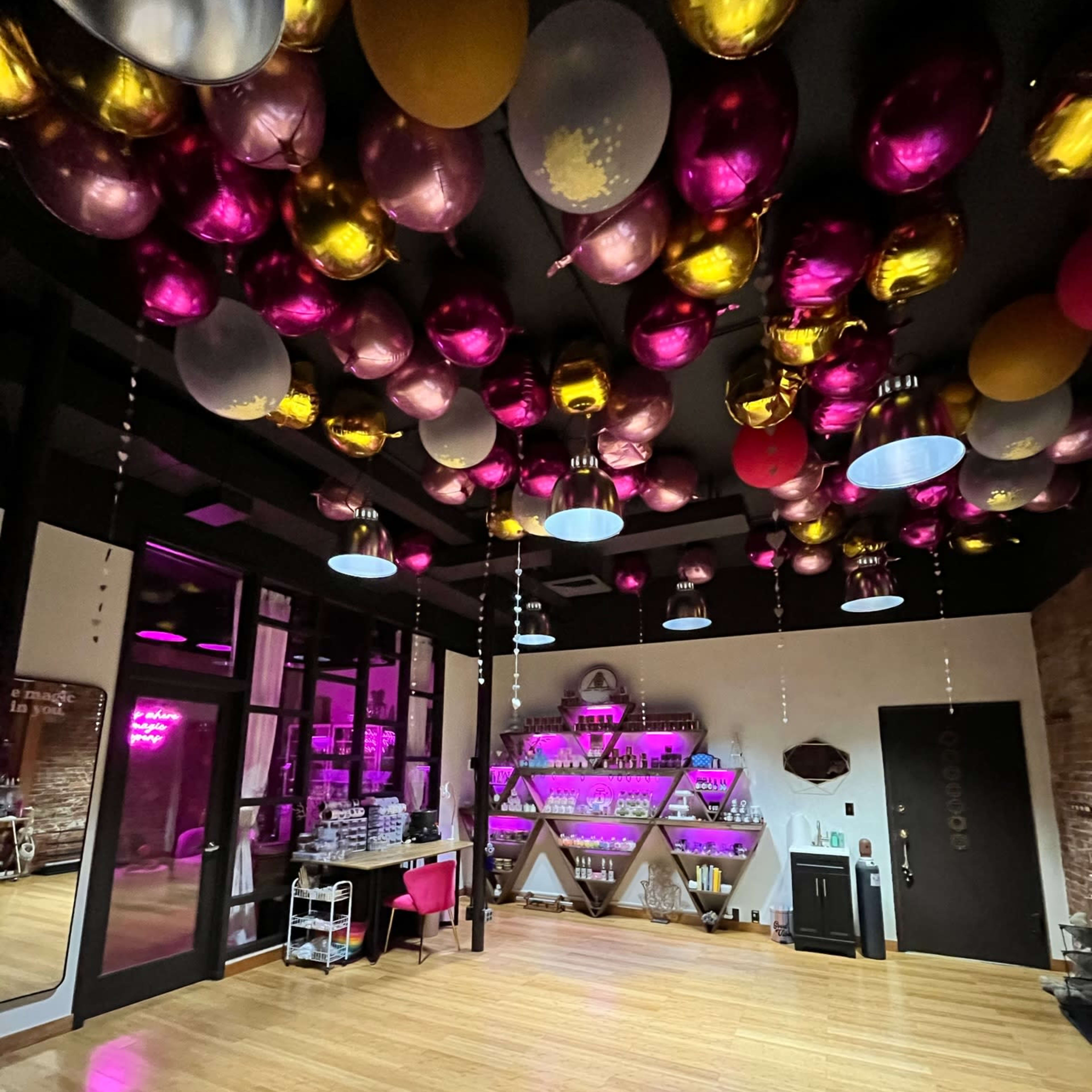 The interior of a venue features a wooden floor, a wall adorned with shelves of colorful bottles, and a ceiling decorated with pink and gold balloons.