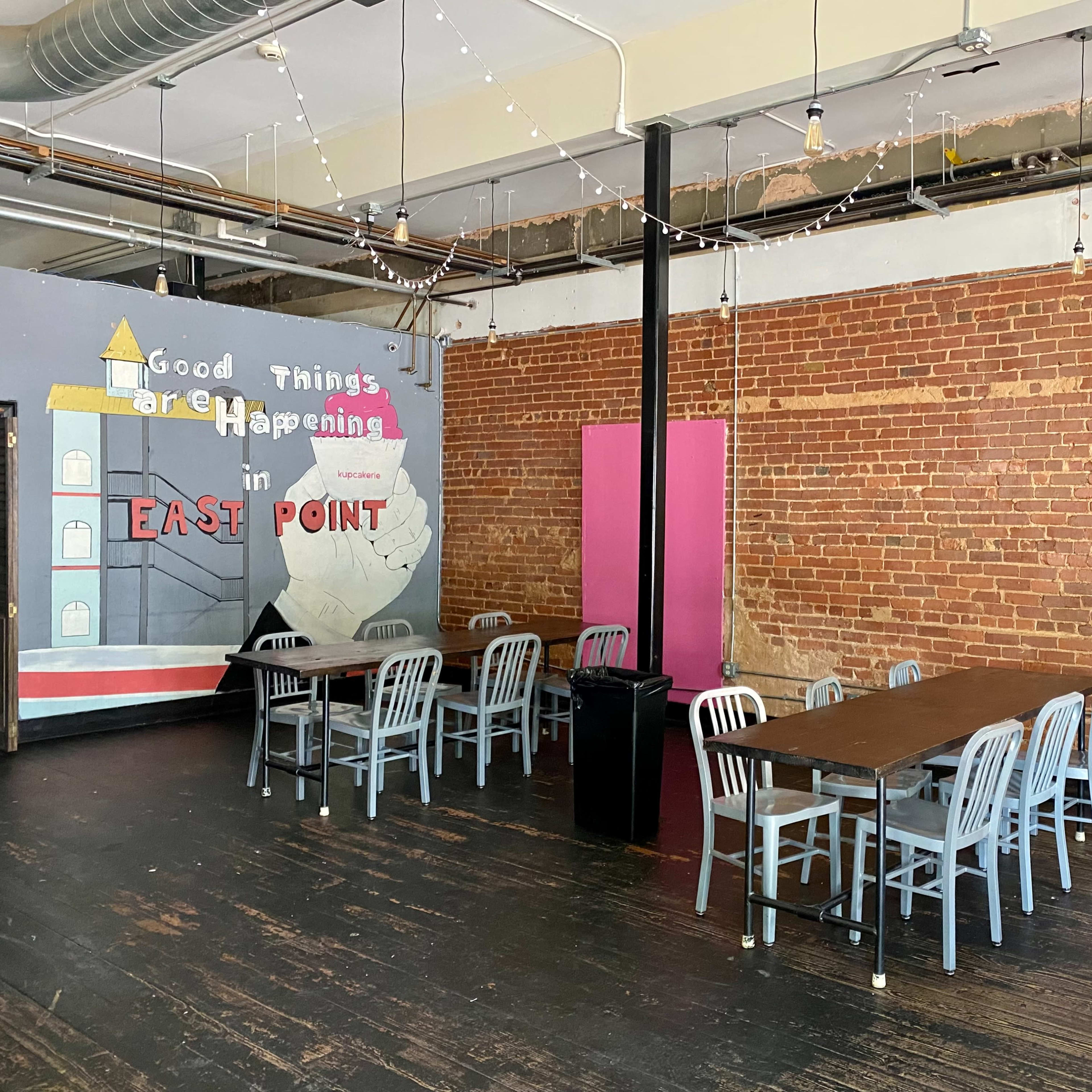 A spacious room with a long table surrounded by chairs, exposed brick walls, and a mural that reads "Good Things Are Happening in East Point."