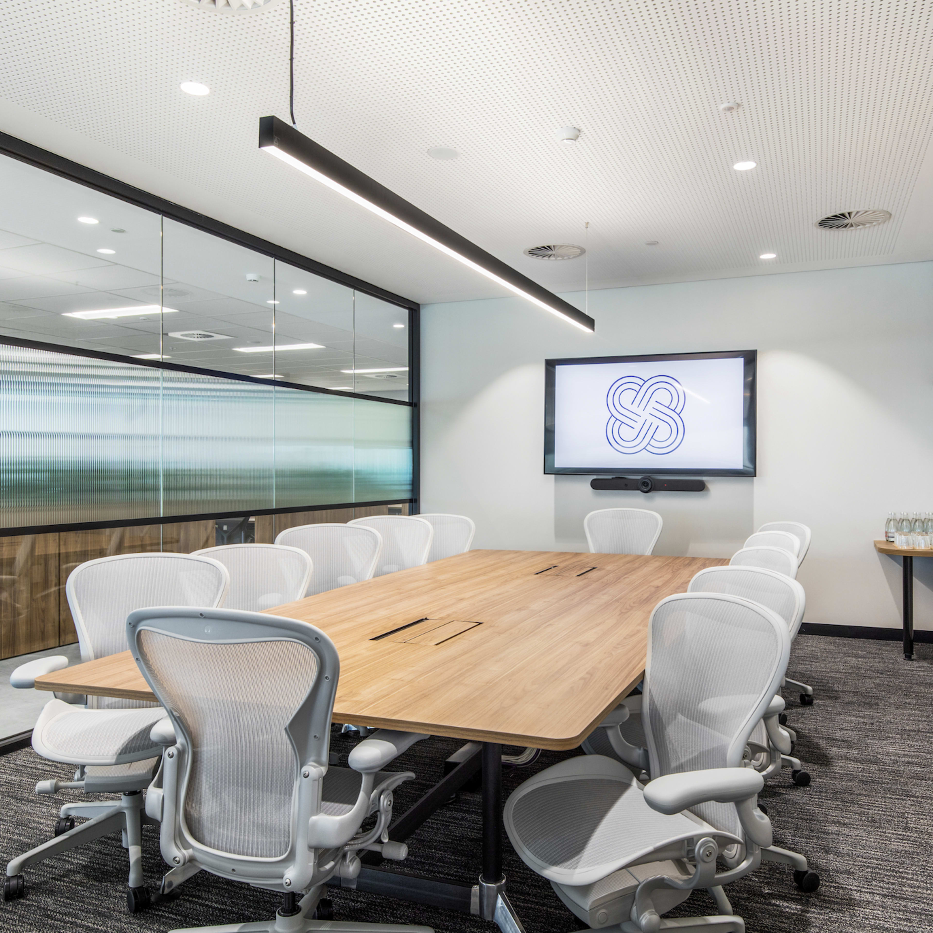 A modern conference room with a long wooden table, white ergonomic chairs, and a wall-mounted display.