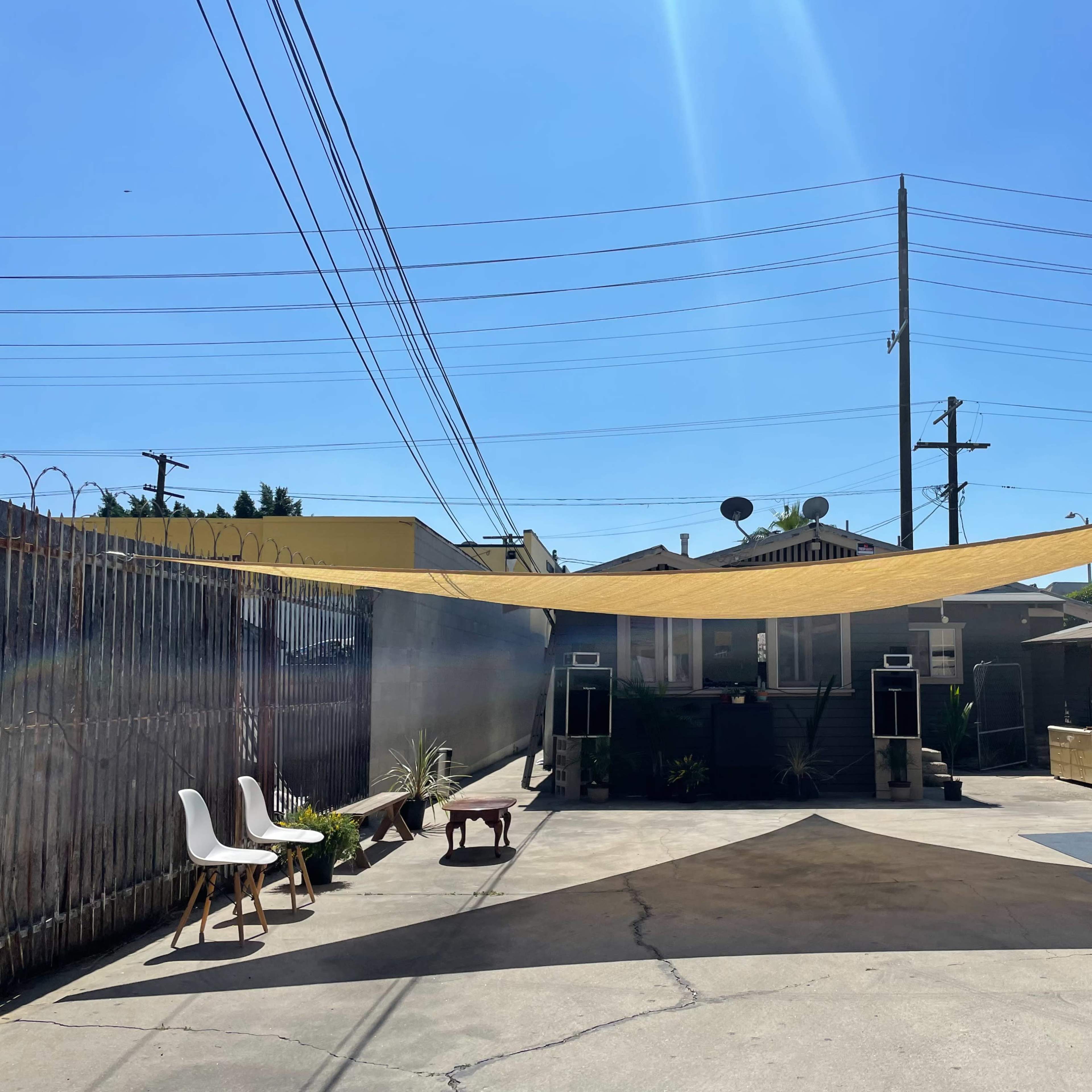 A large yellow shade sail stretches across a concrete yard, with a few chairs and plants arranged below, surrounded by buildings and power lines.