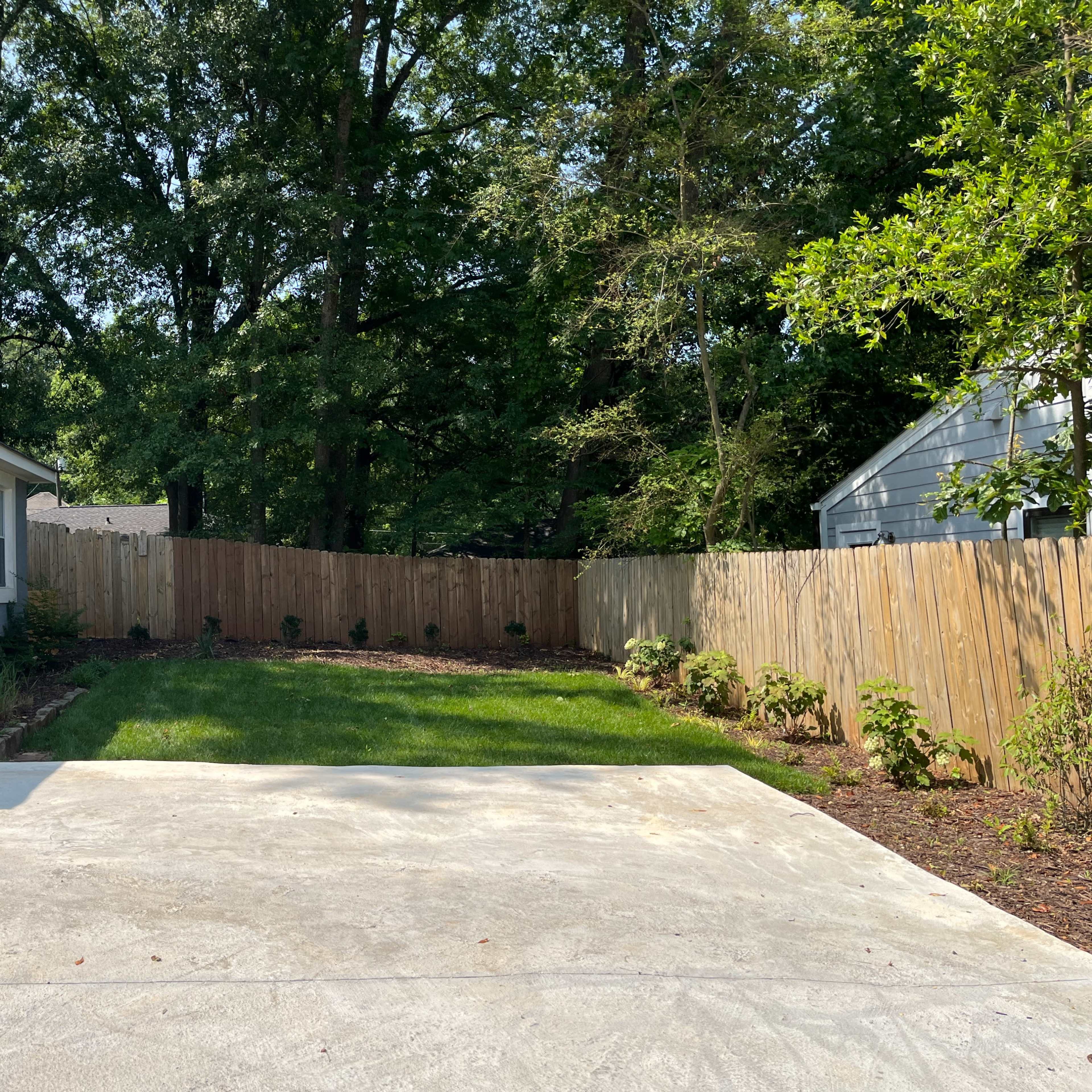 A clear, fenced backyard with a small grassy area and two houses visible on either side.