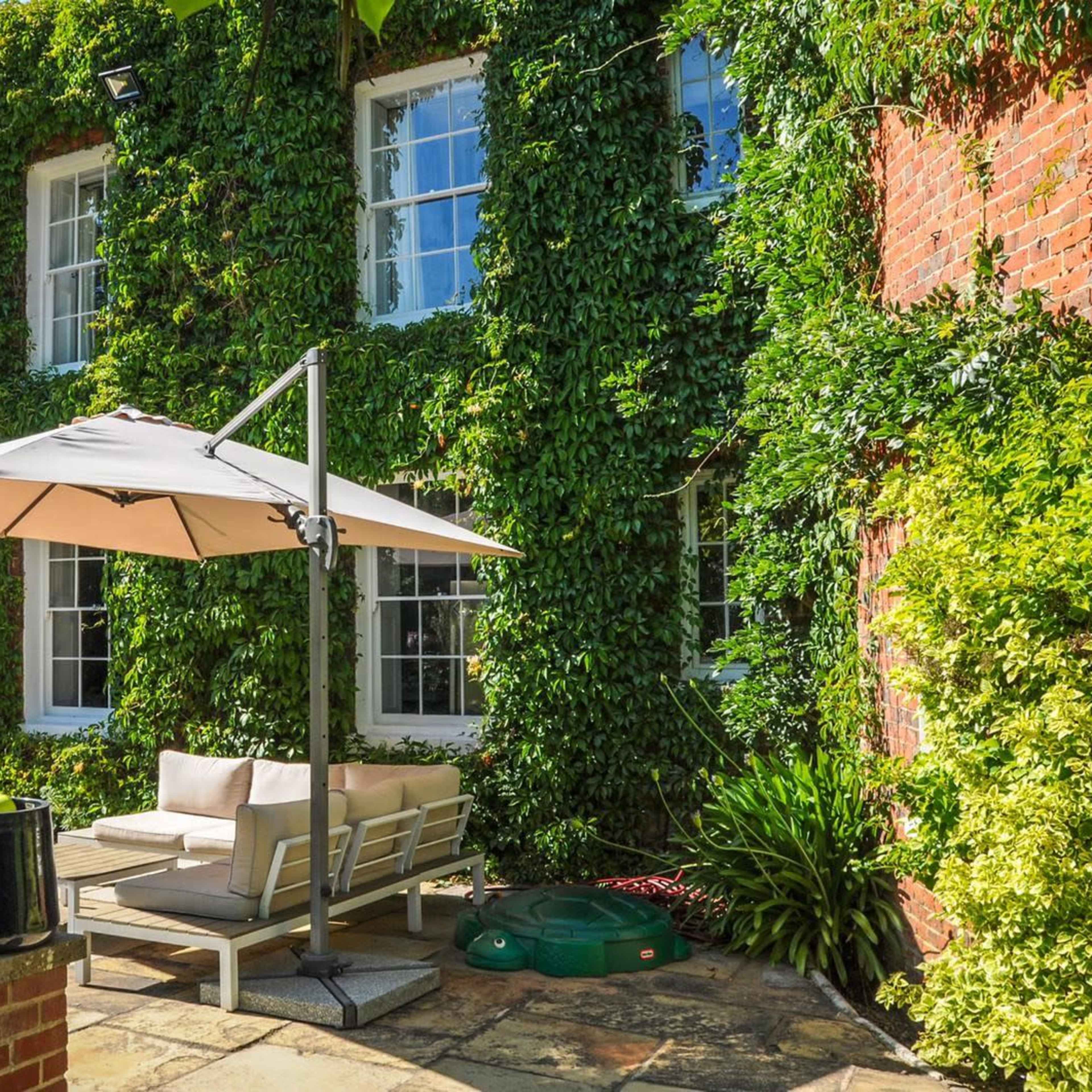 A shaded seating area with a light umbrella and sofa is surrounded by a lush, green vine-covered wall and brick facade.