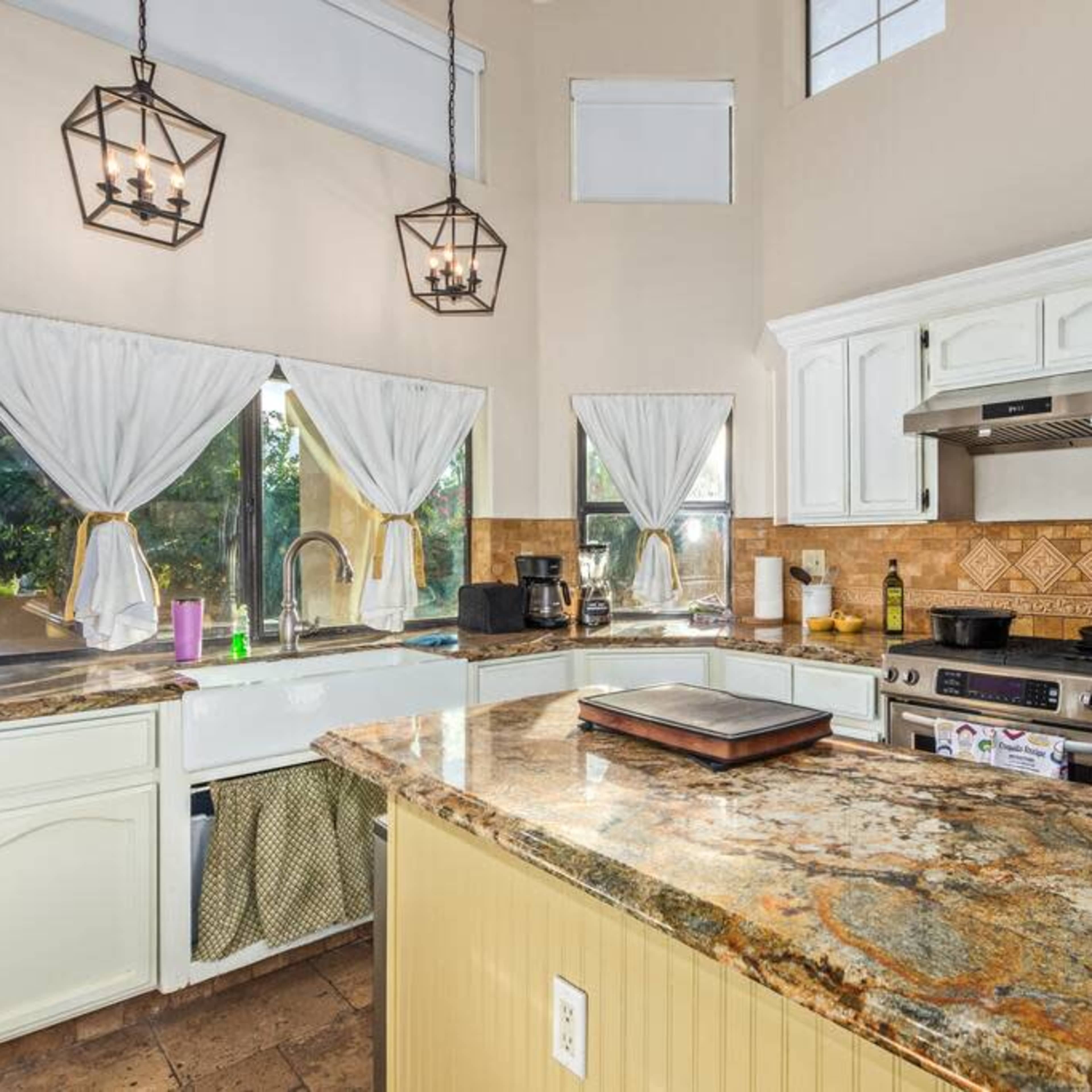 A modern kitchen features white cabinetry, granite countertops, and large windows with sheer curtains.