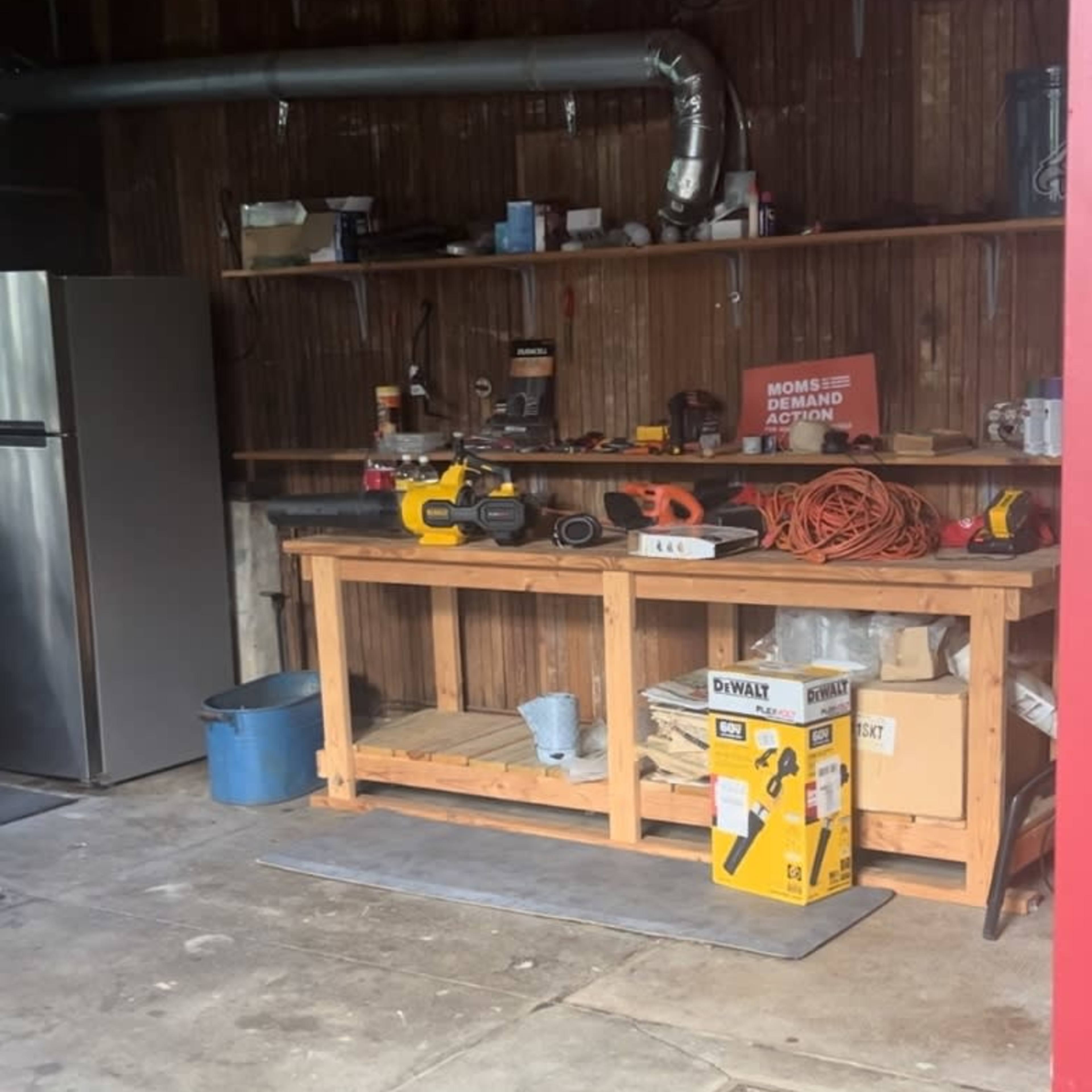 A cluttered garage workshop with a workbench, various tools, a refrigerator, and a blue bin.