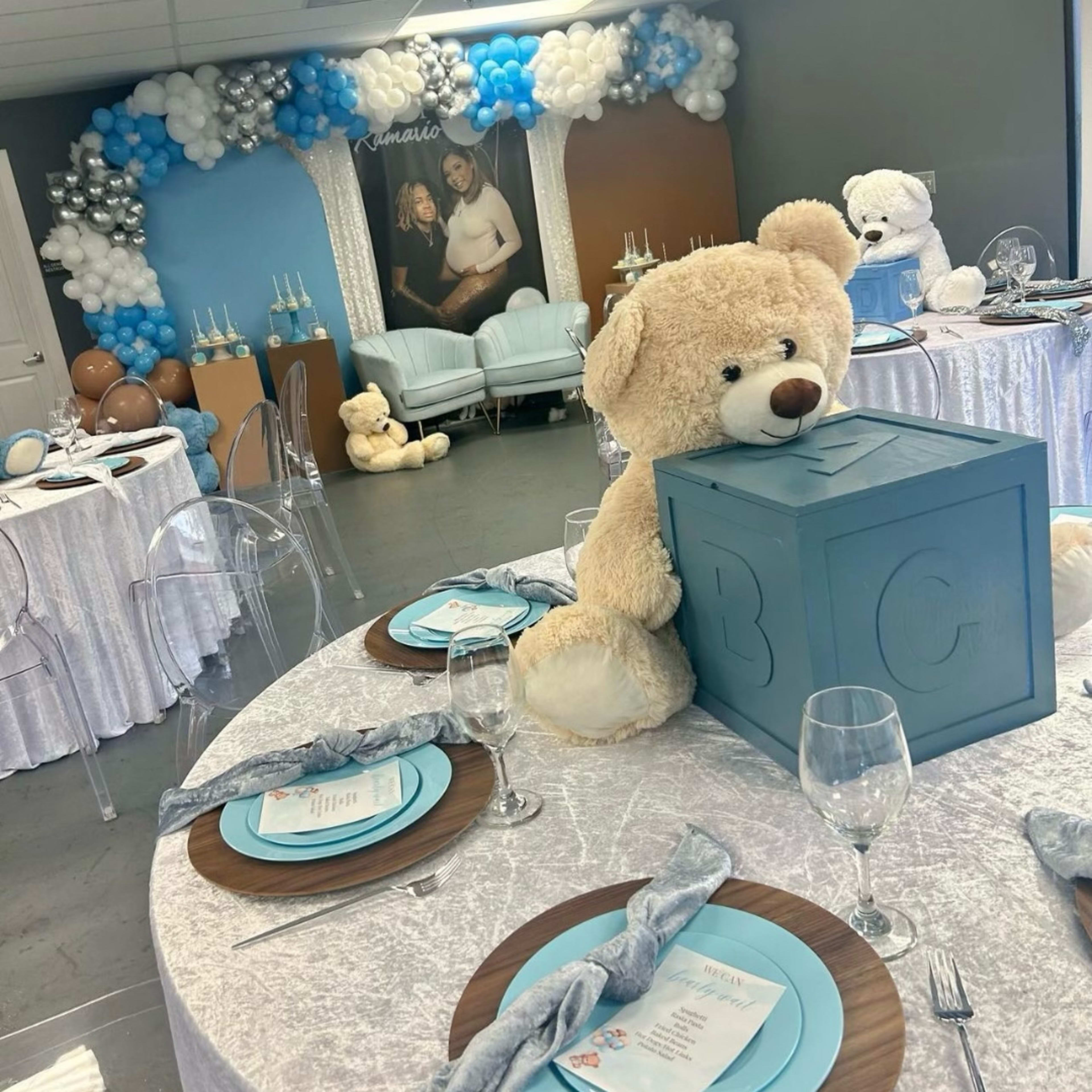 A decorated event space with tables set for a celebration, featuring large teddy bears, balloons, and a backdrop with a photo of a couple.