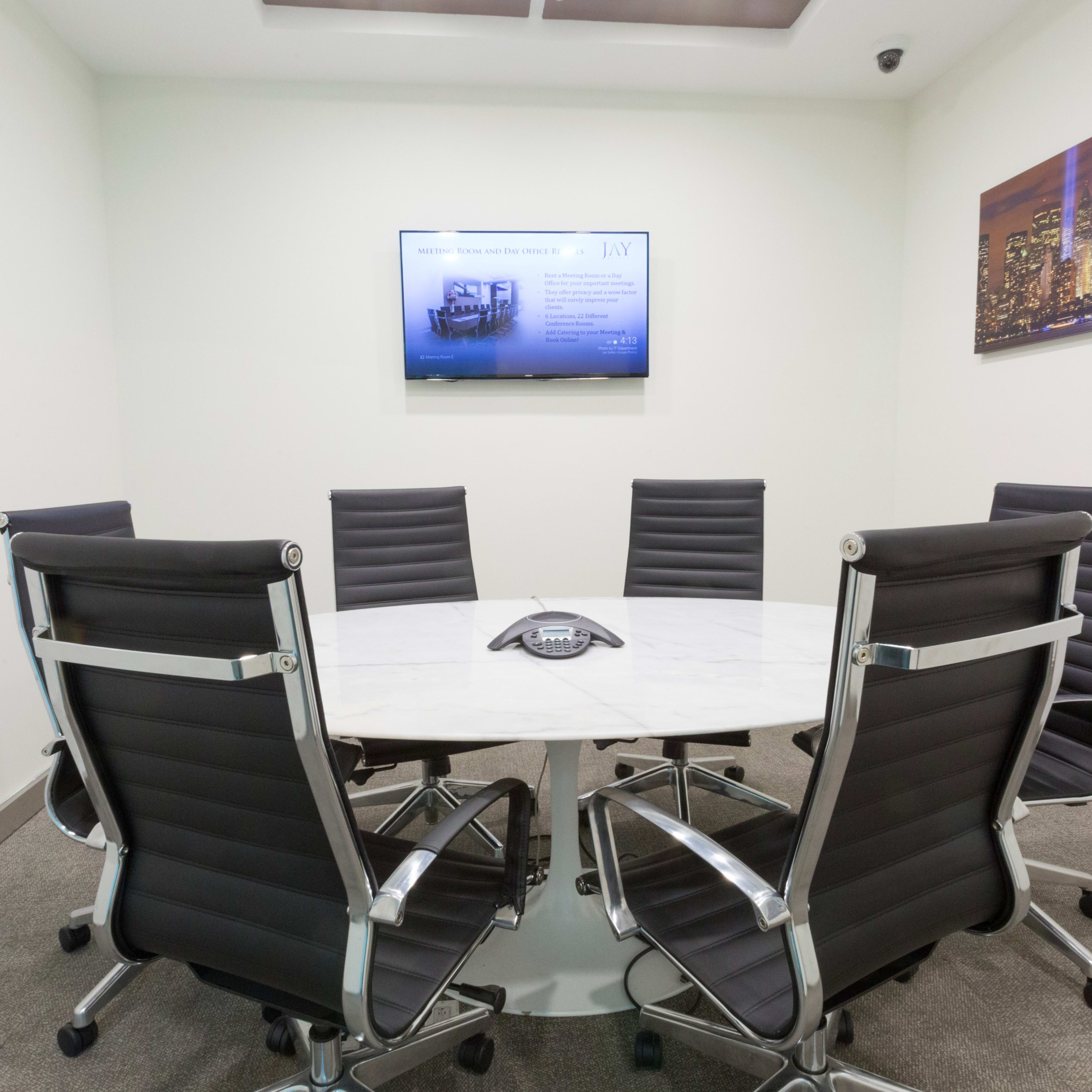 A modern conference room features a round table surrounded by six black office chairs and a wall-mounted television displaying a presentation.