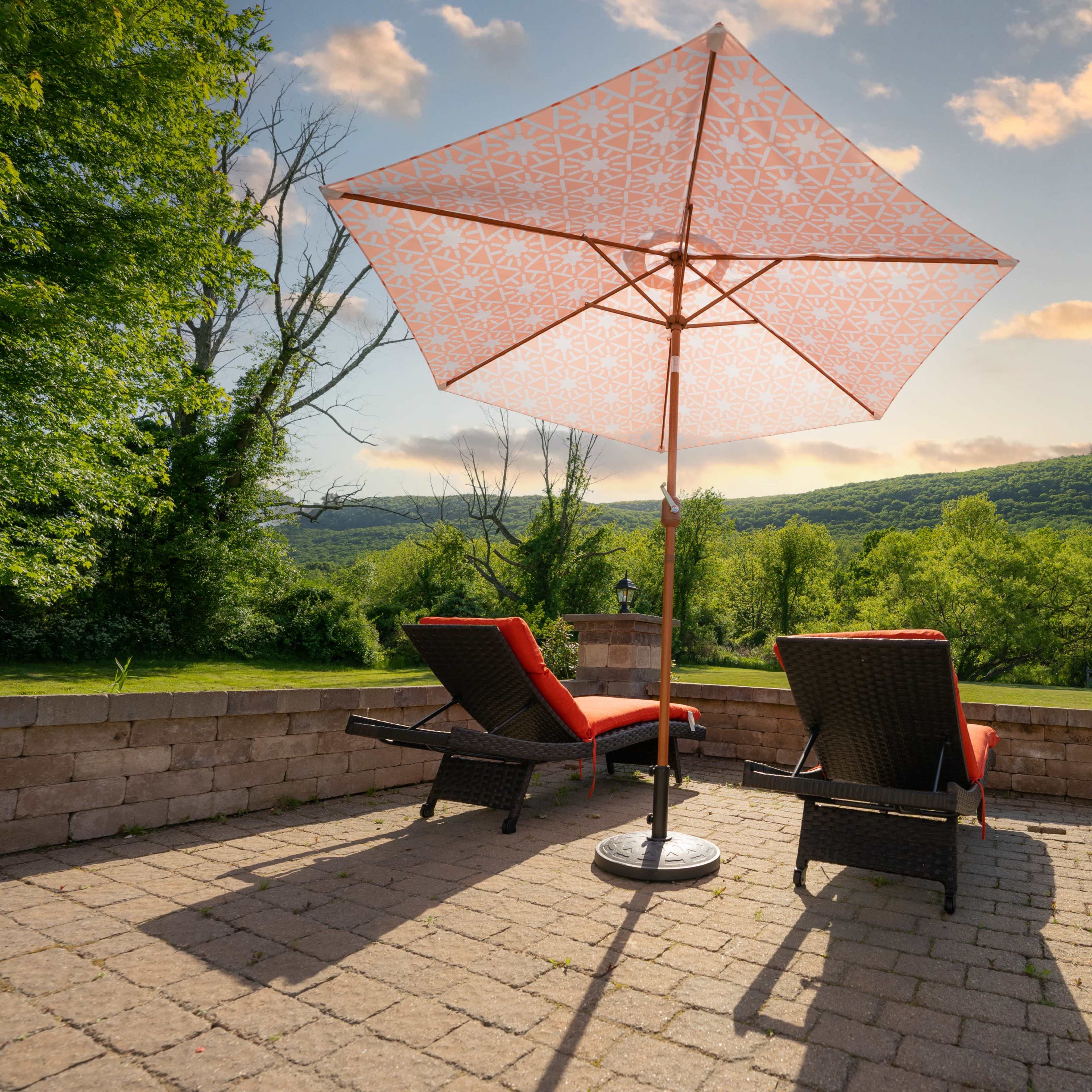 Two lounge chairs with red cushions are positioned under a patterned umbrella, overlooking a green landscape and a cloudy sky.