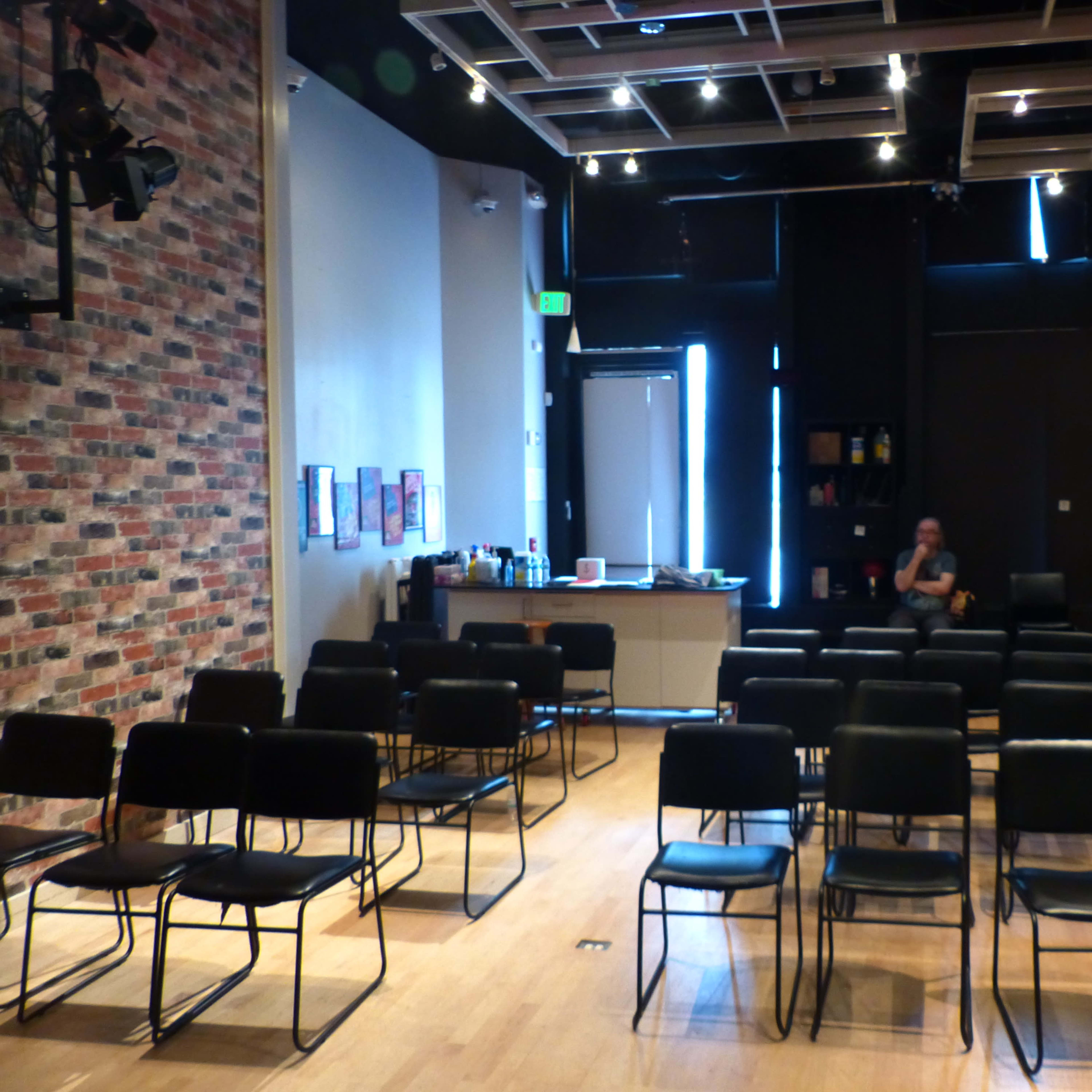 A nearly empty room with rows of chairs facing a stage, featuring a brick wall, and a lone person seated in the back.
