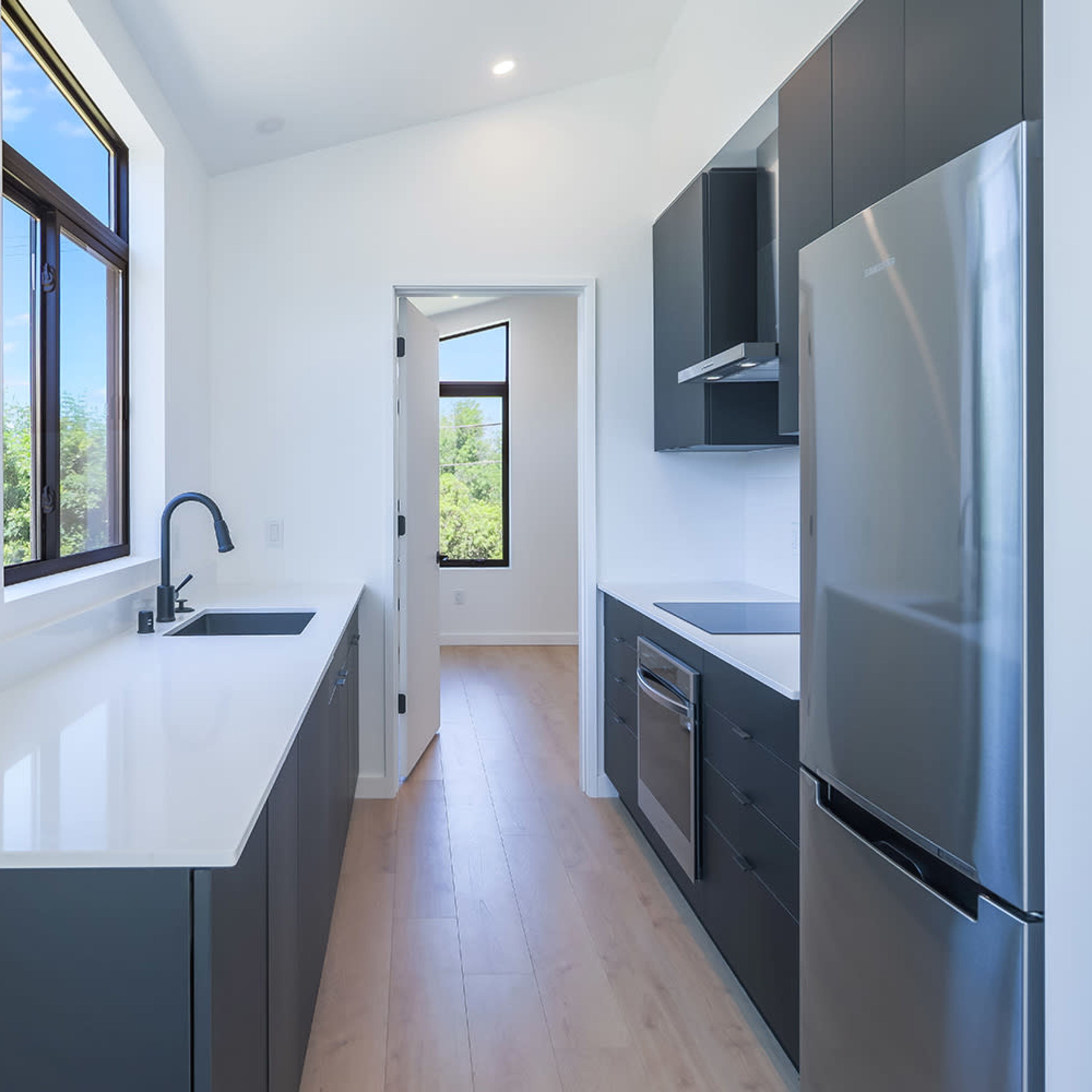 A narrow kitchen features sleek cabinetry, a stainless steel refrigerator, and a large window allowing natural light.