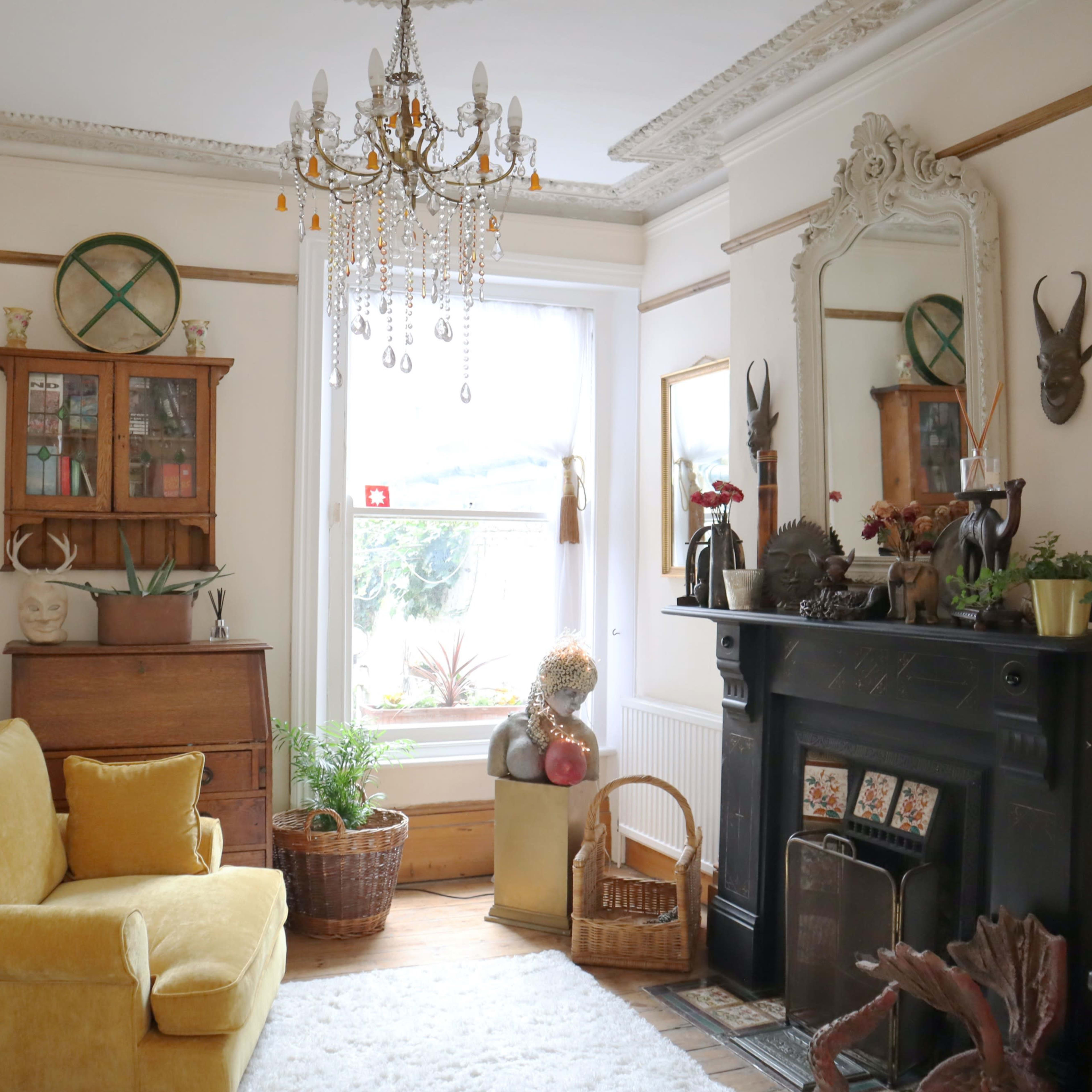 A cozy living room features a yellow couch, a wooden cabinet, a decorative mirror, and a fireplace surrounded by plants and eclectic decor.