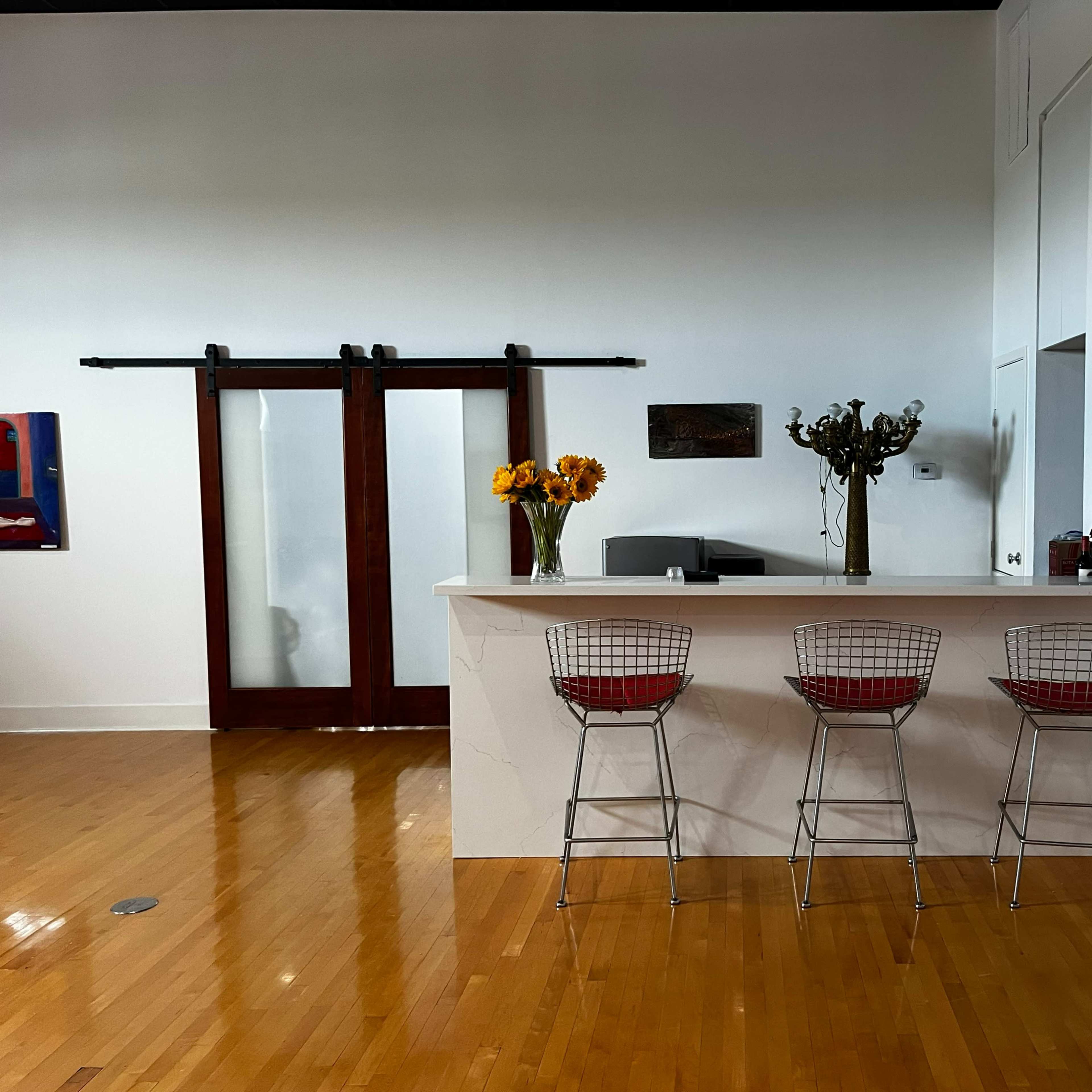 A modern kitchen bar with three wire stools sits in front of a counter, flanked by artwork and a sliding glass door.