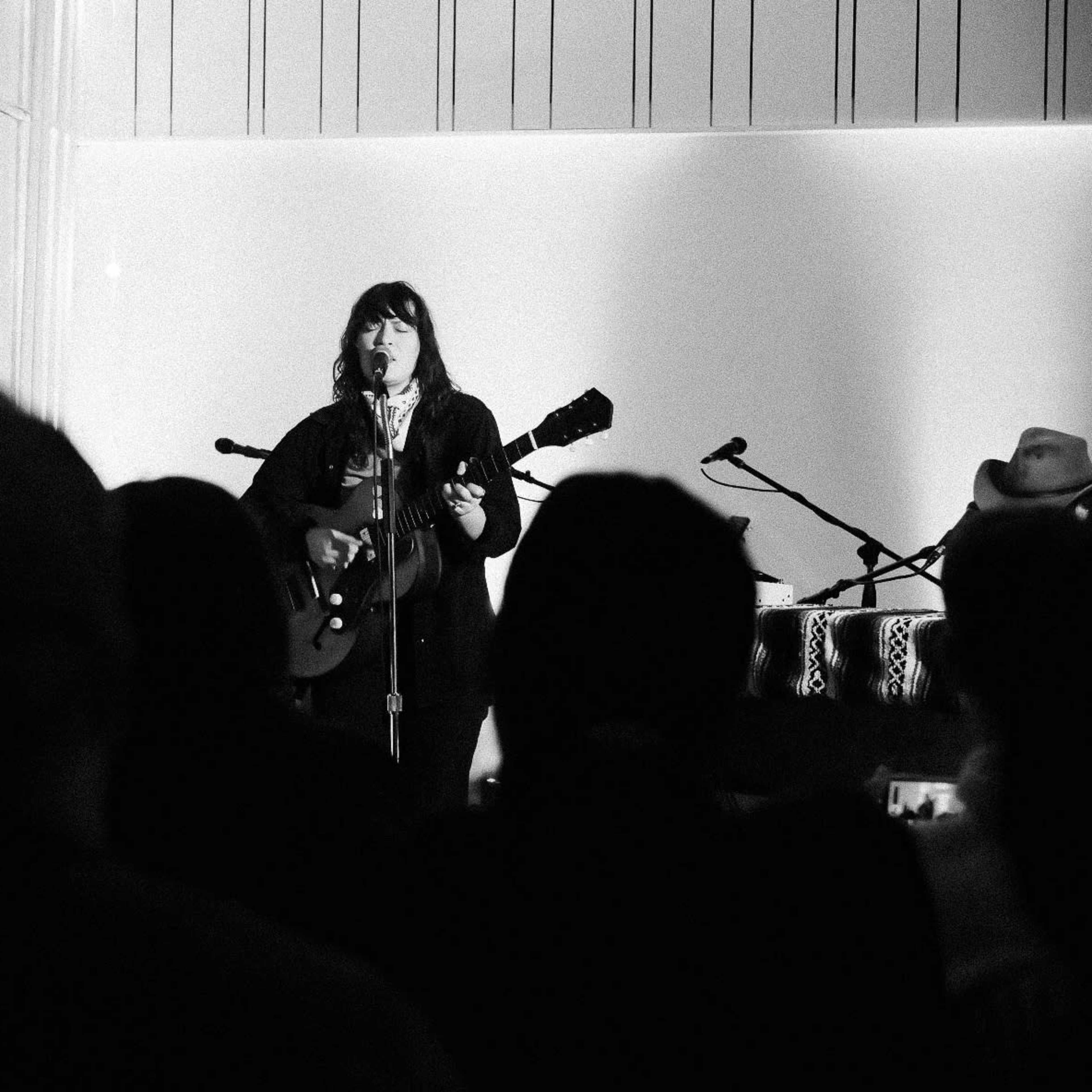 A musician stands on stage, playing a guitar while an audience watches from the foreground.