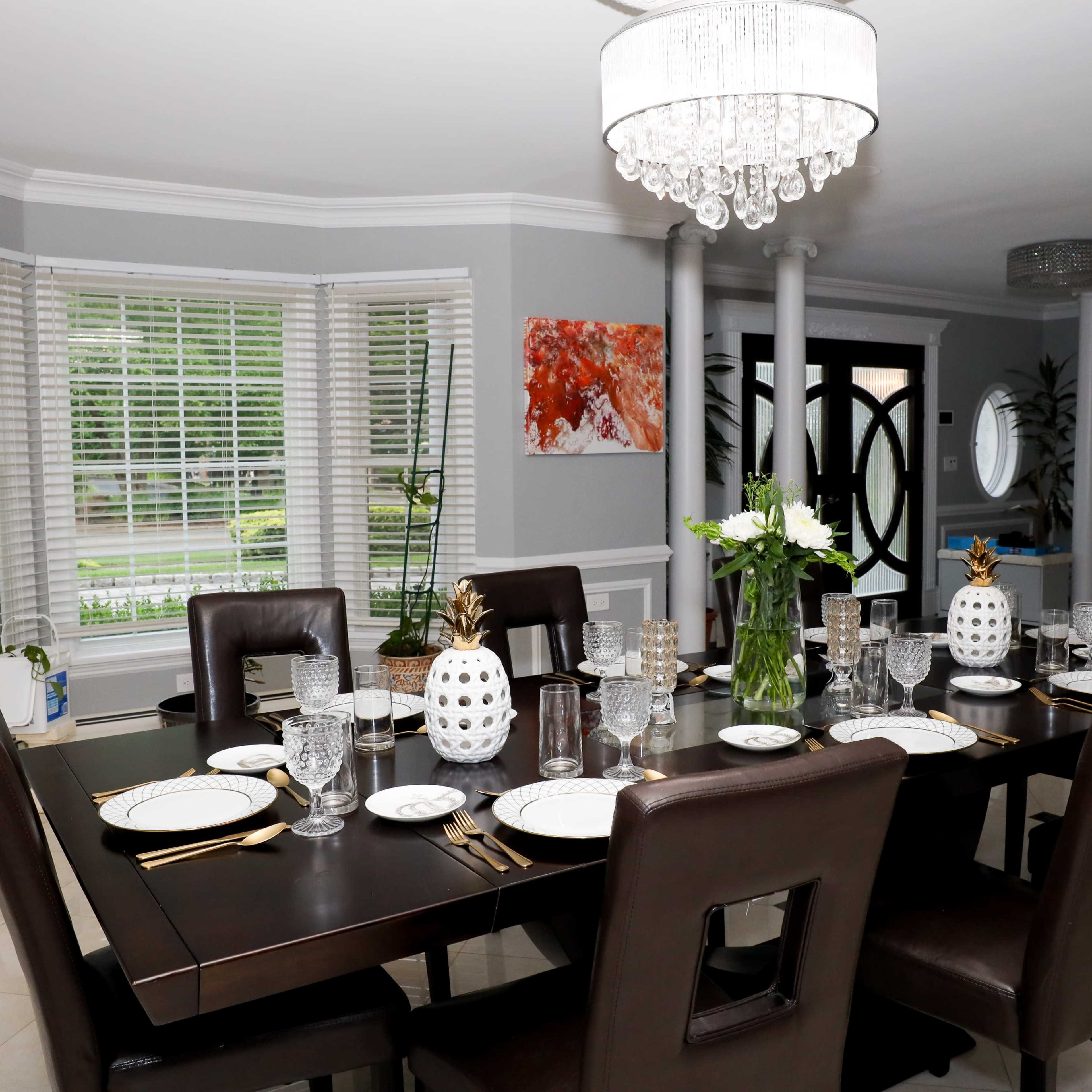 A formal dining room features a long dark table set with plates, glasses, and decorative centerpieces, surrounded by brown chairs under a crystal chandelier.
