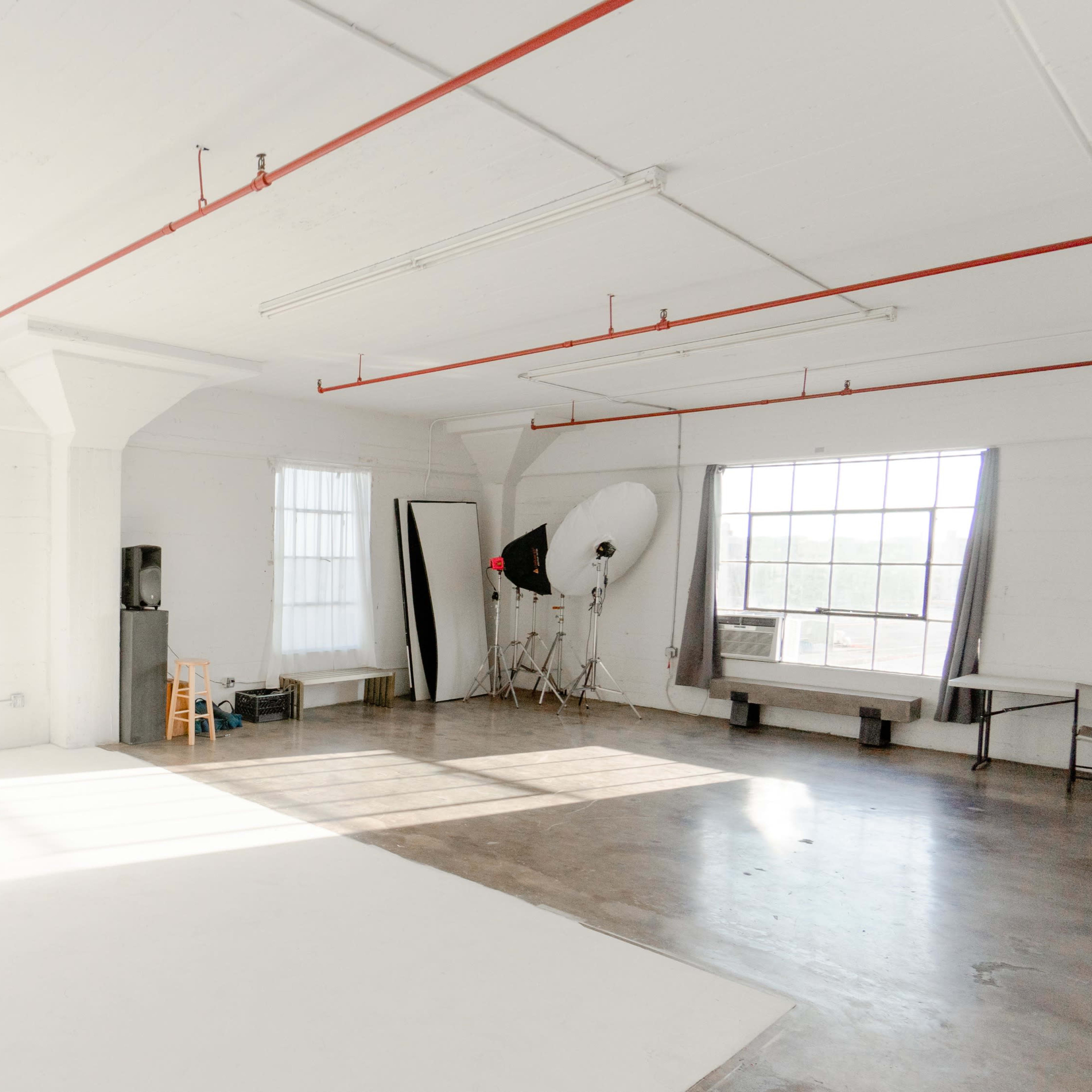 A spacious, well-lit photography studio featuring white walls, large windows, and various studio equipment.