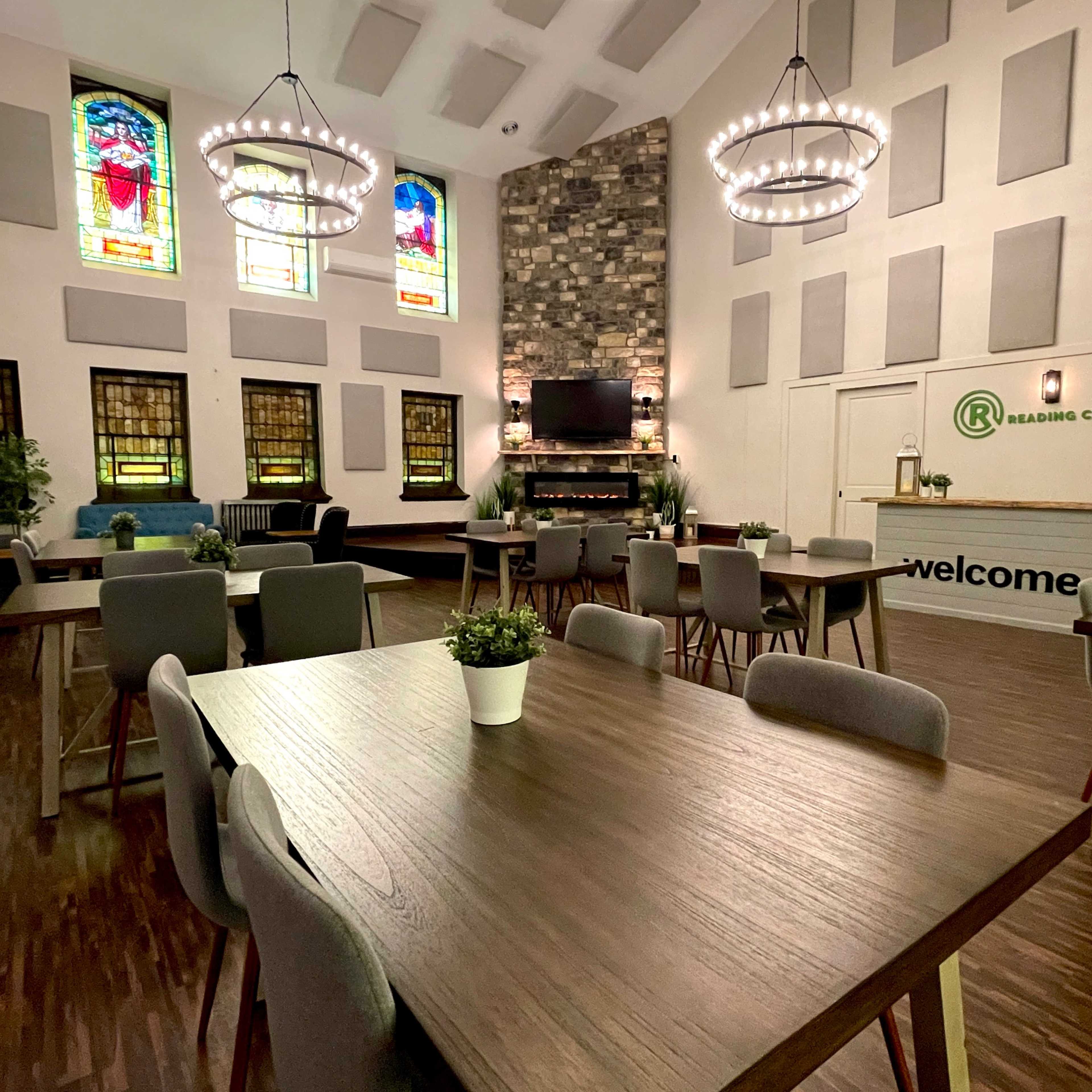 The interior of a church features tables and chairs set up for gatherings, with stained glass windows and a stone fireplace as focal points.
