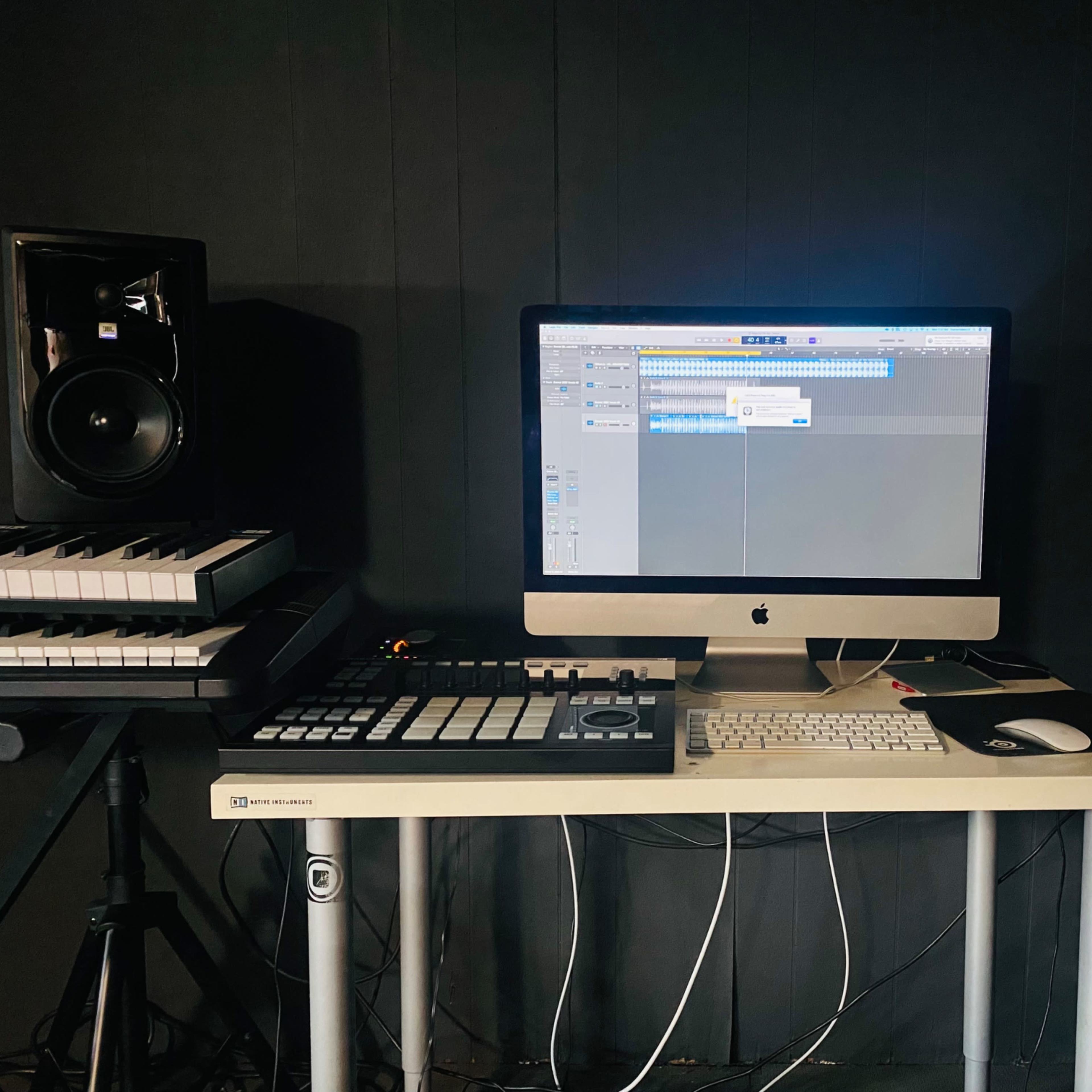 A music production workspace featuring an iMac with a digital audio workstation open, flanked by studio monitors and a MIDI controller on a desk.