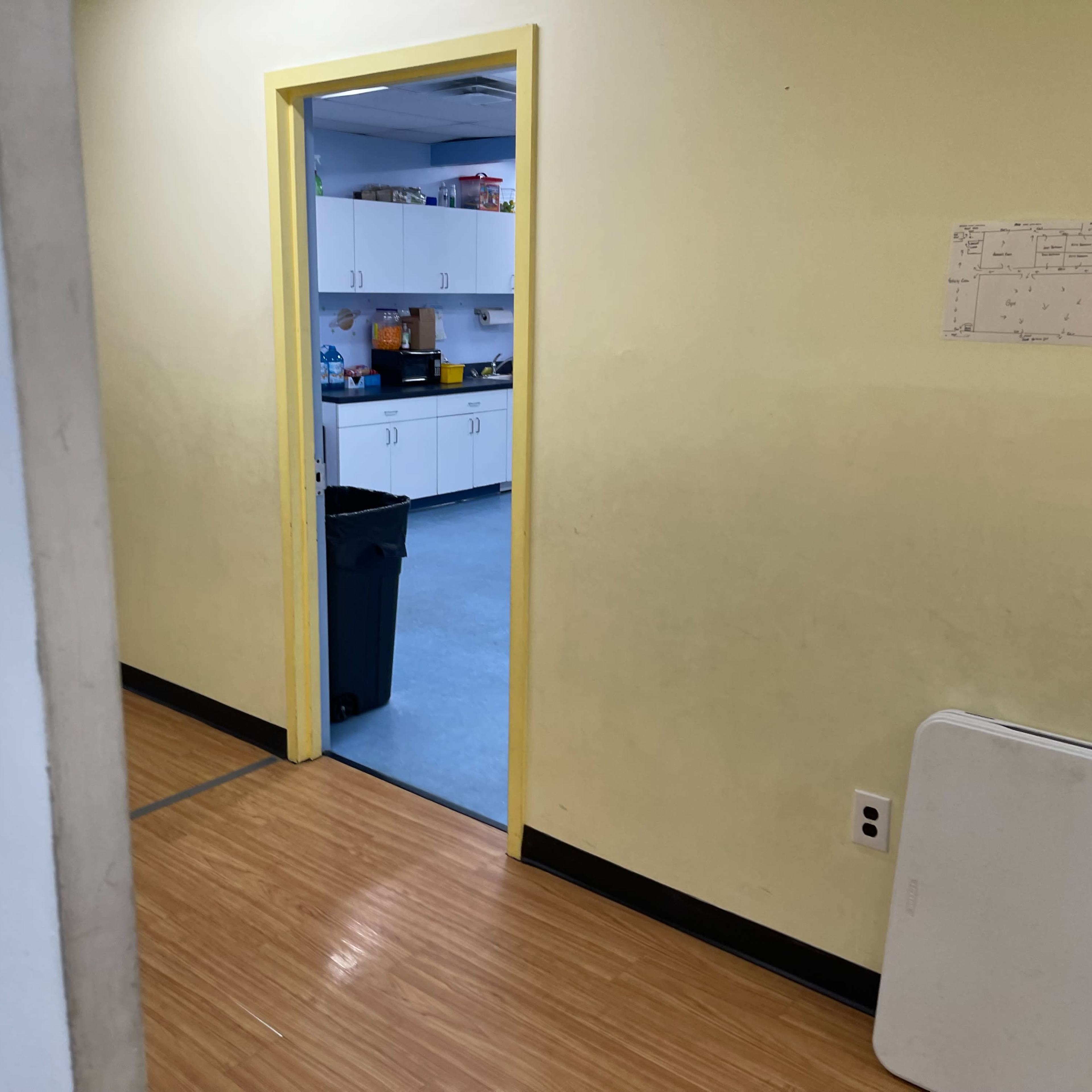 A yellow-walled corridor leads to a kitchen area with white cabinets and a black trash can.