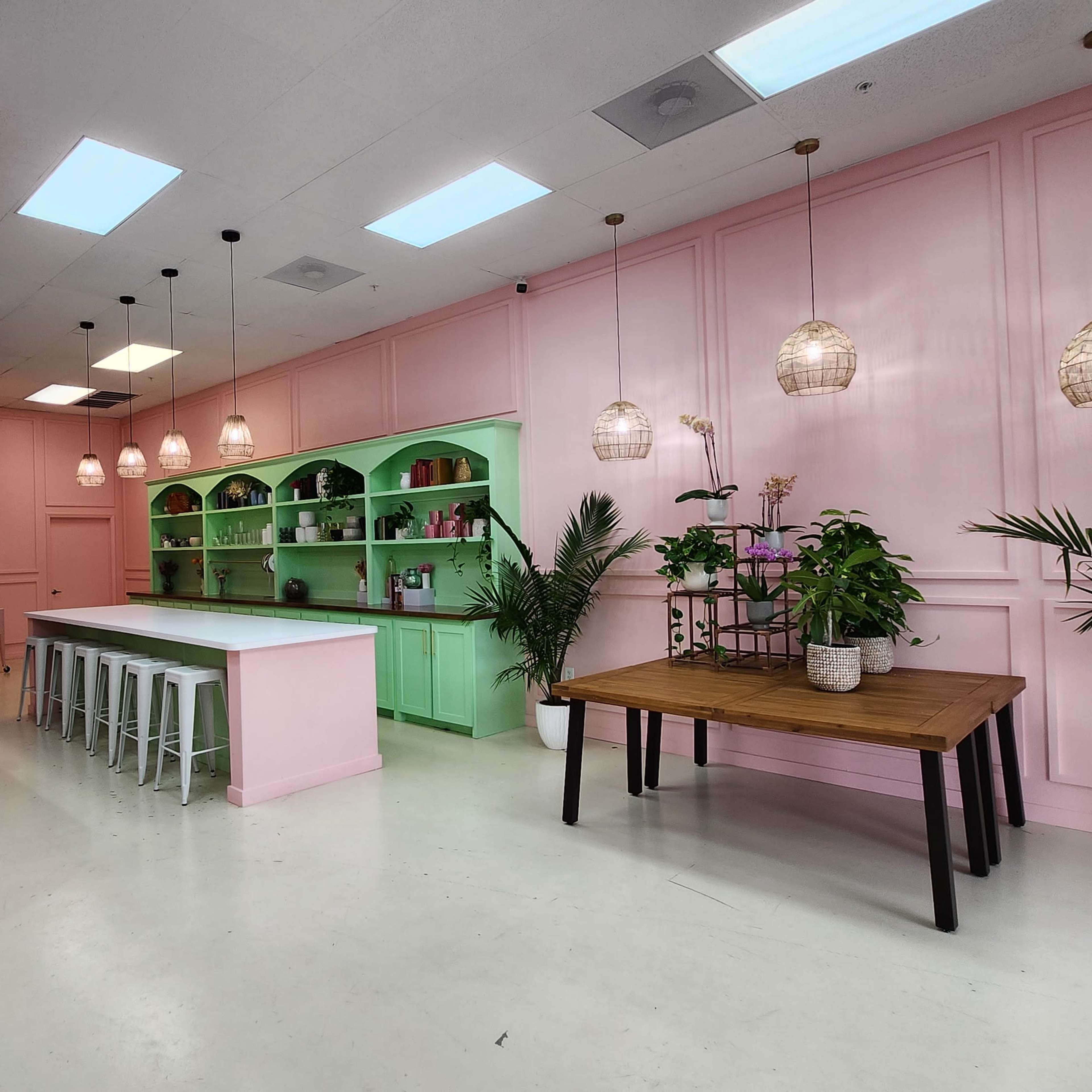 A brightly lit interior space featuring pink and green walls, a central island with white stools, and several plants and hanging lights.