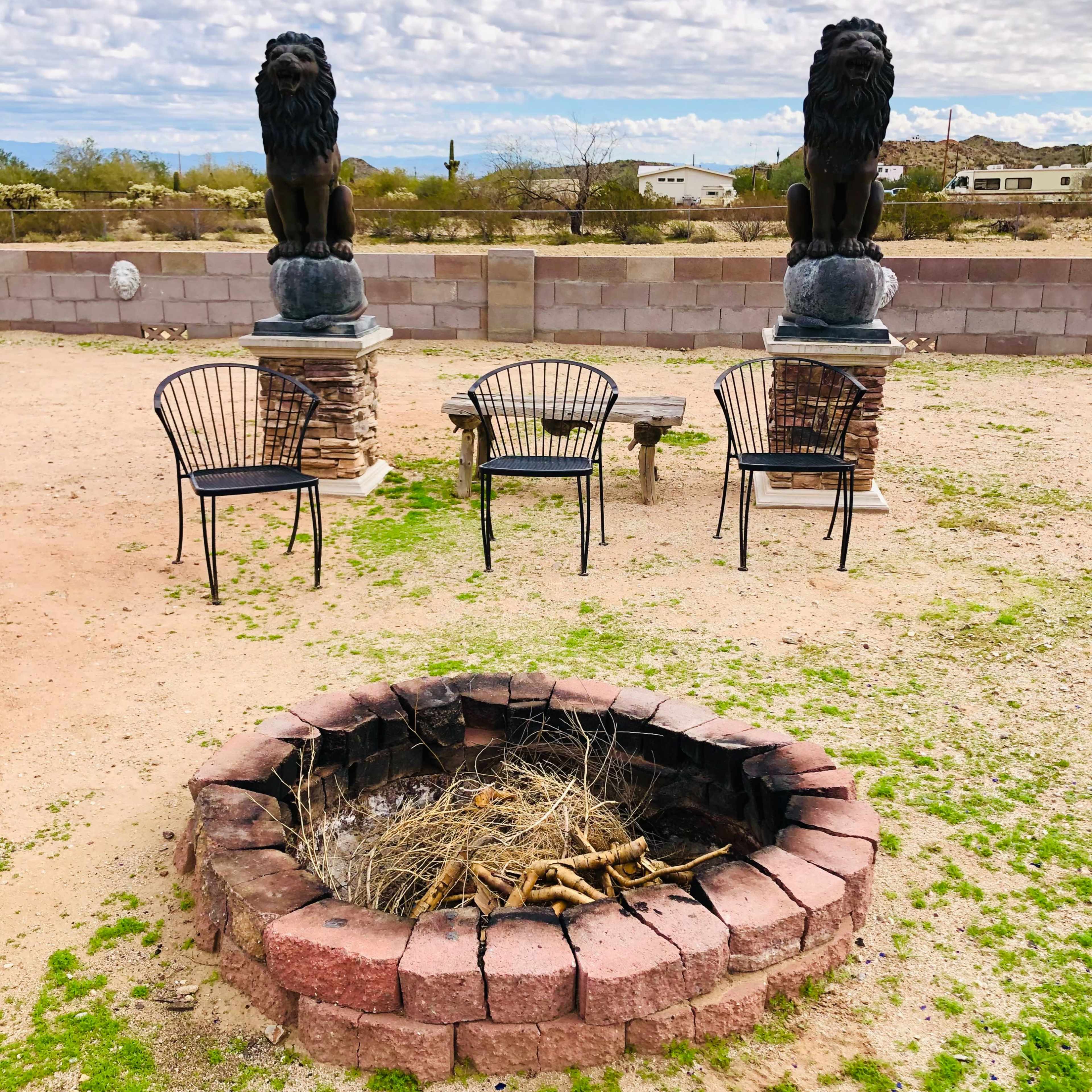 A circular stone fire pit surrounded by three black chairs, with two lion statues on raised pedestals in the background.