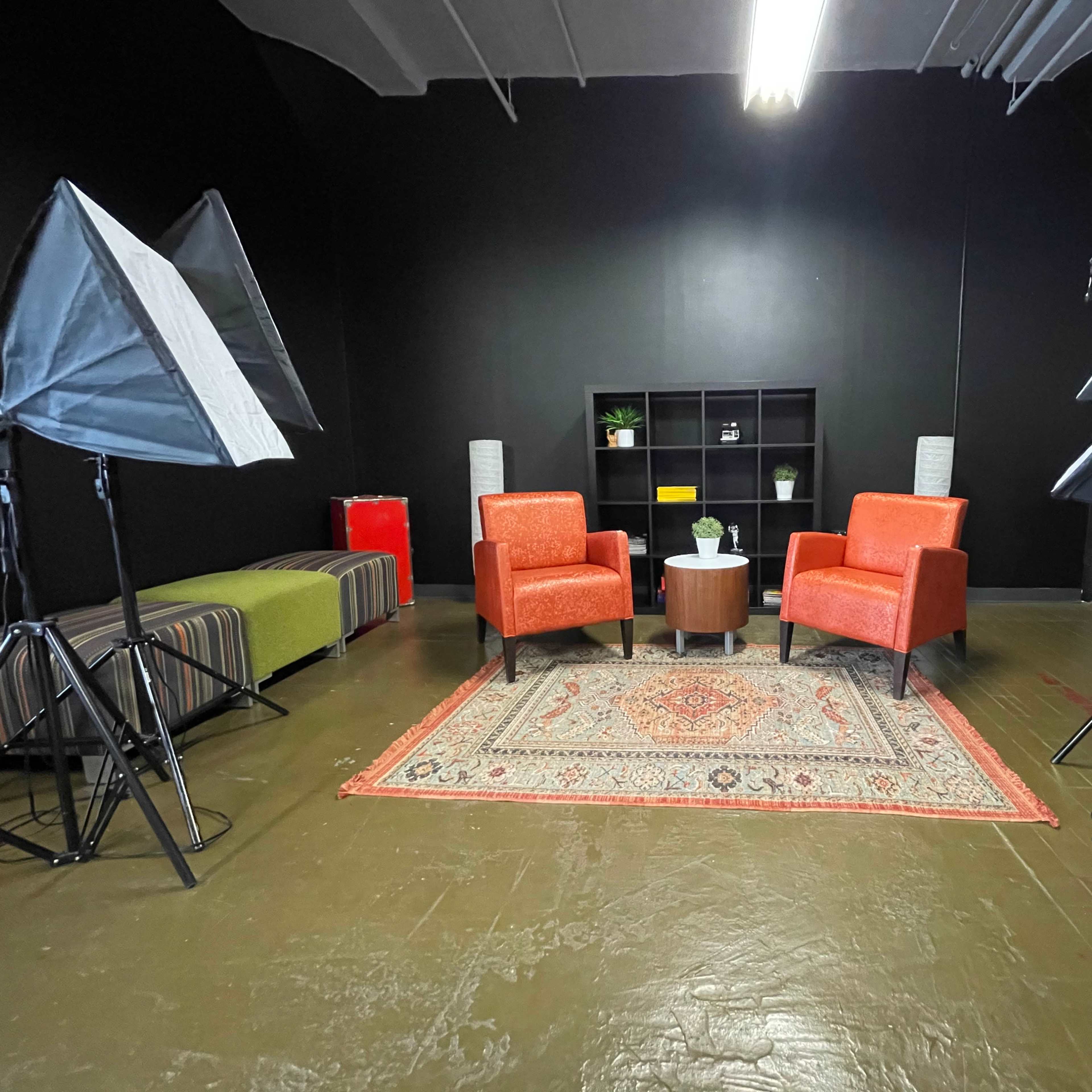 A studio setup featuring two orange chairs, a small round table, and multiple softbox lights arranged around a patterned rug on a dark wall background.