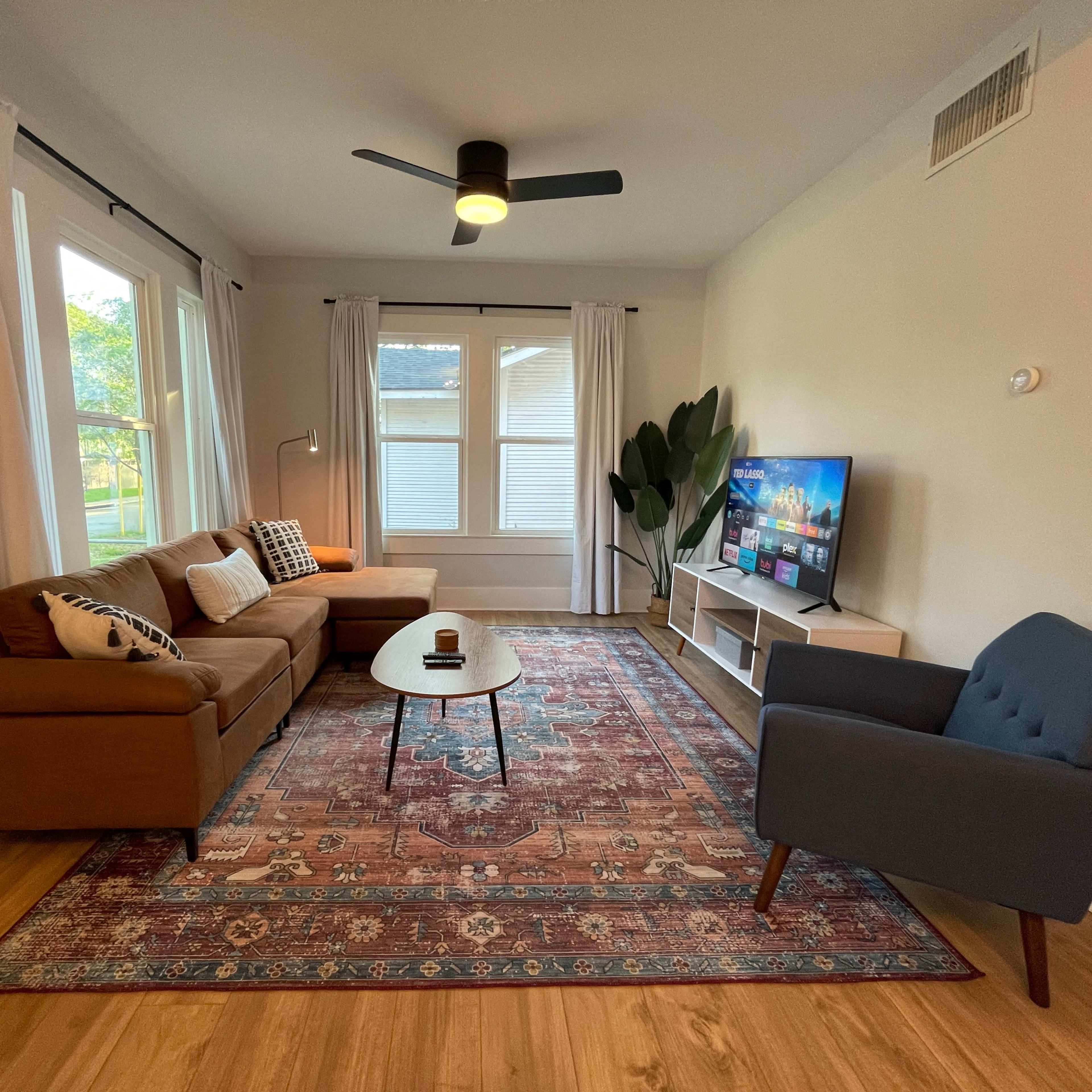 The living room features a brown sofa, a blue armchair, a round coffee table, and a television, all arranged around a patterned rug under a ceiling fan and natural light from two windows.