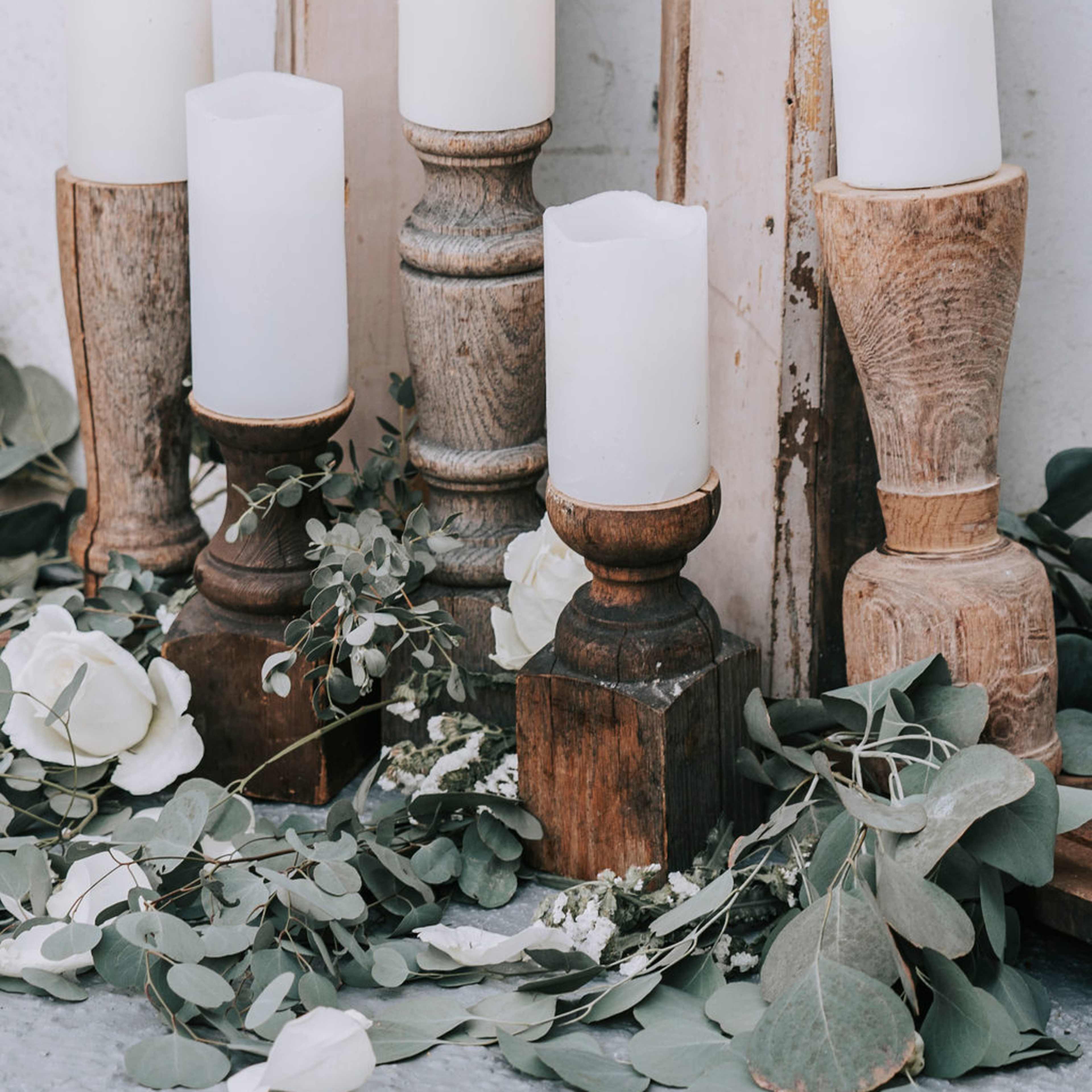 A collection of five white candles on wooden candle holders is arranged among greenery and white roses.