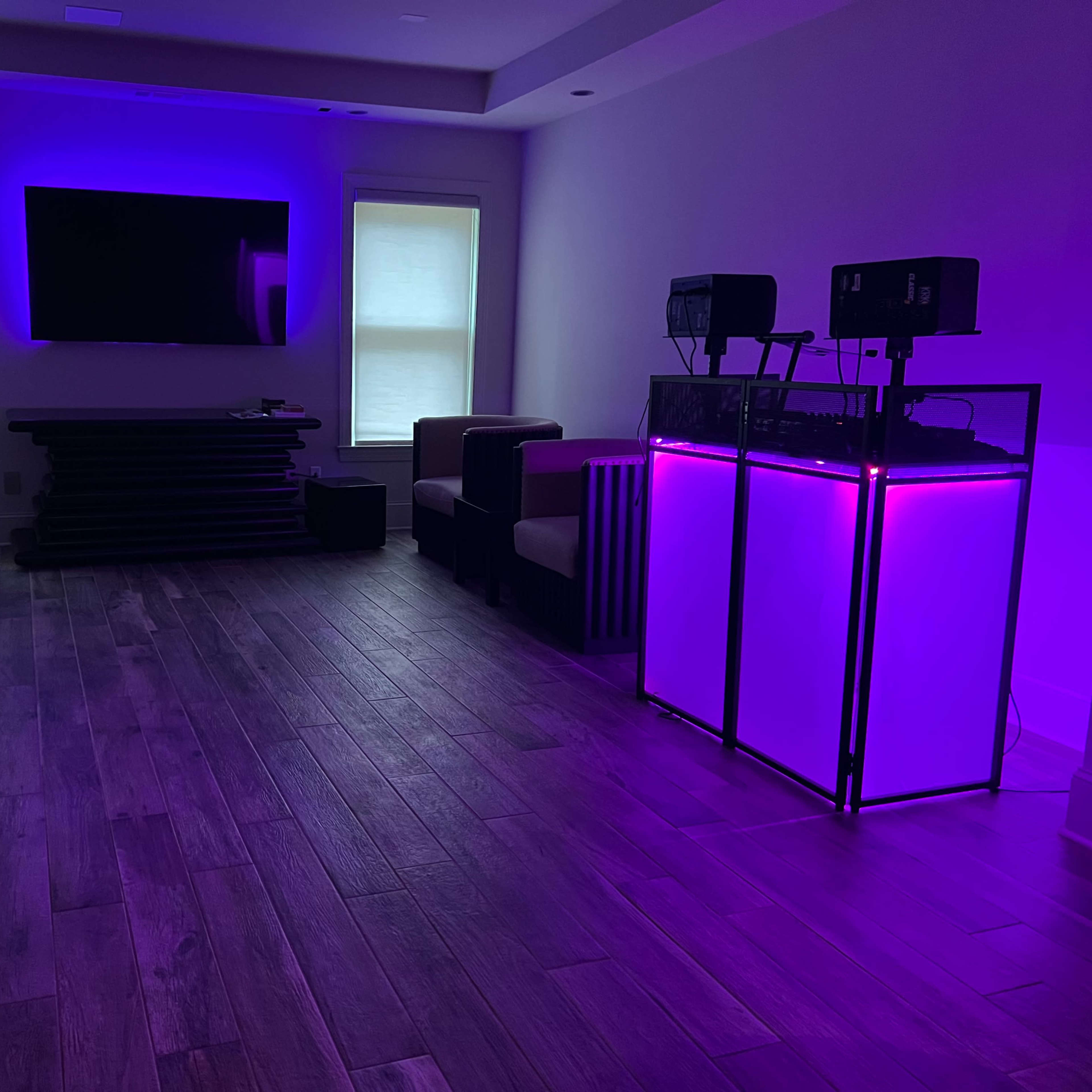 The room features a modern entertainment area with a large TV, two couches, and a DJ setup illuminated by purple lighting.