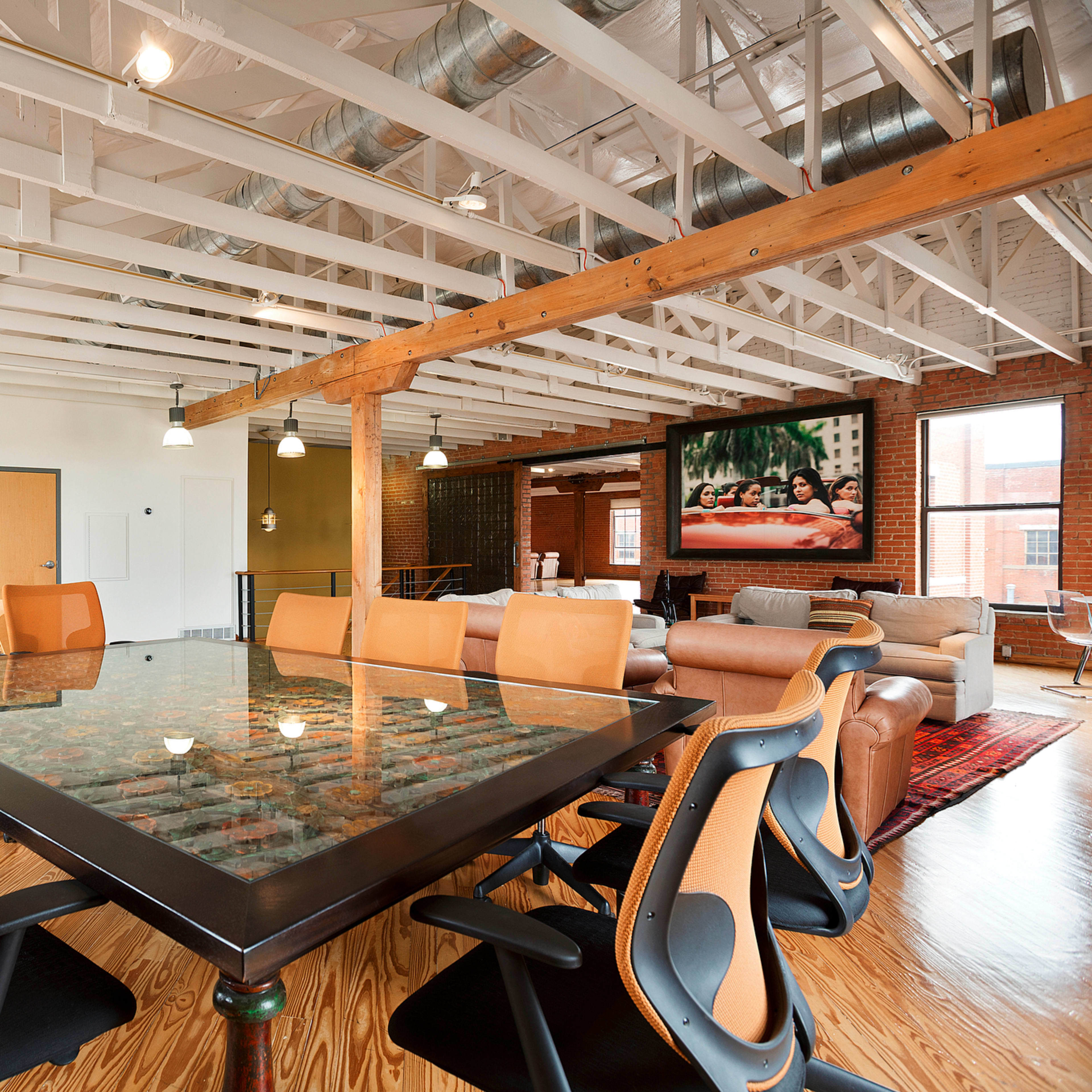 A modern office space with a large glass conference table surrounded by ergonomic chairs, hardwood flooring, and exposed beams, featuring a lounge area with couches and artwork on the walls.