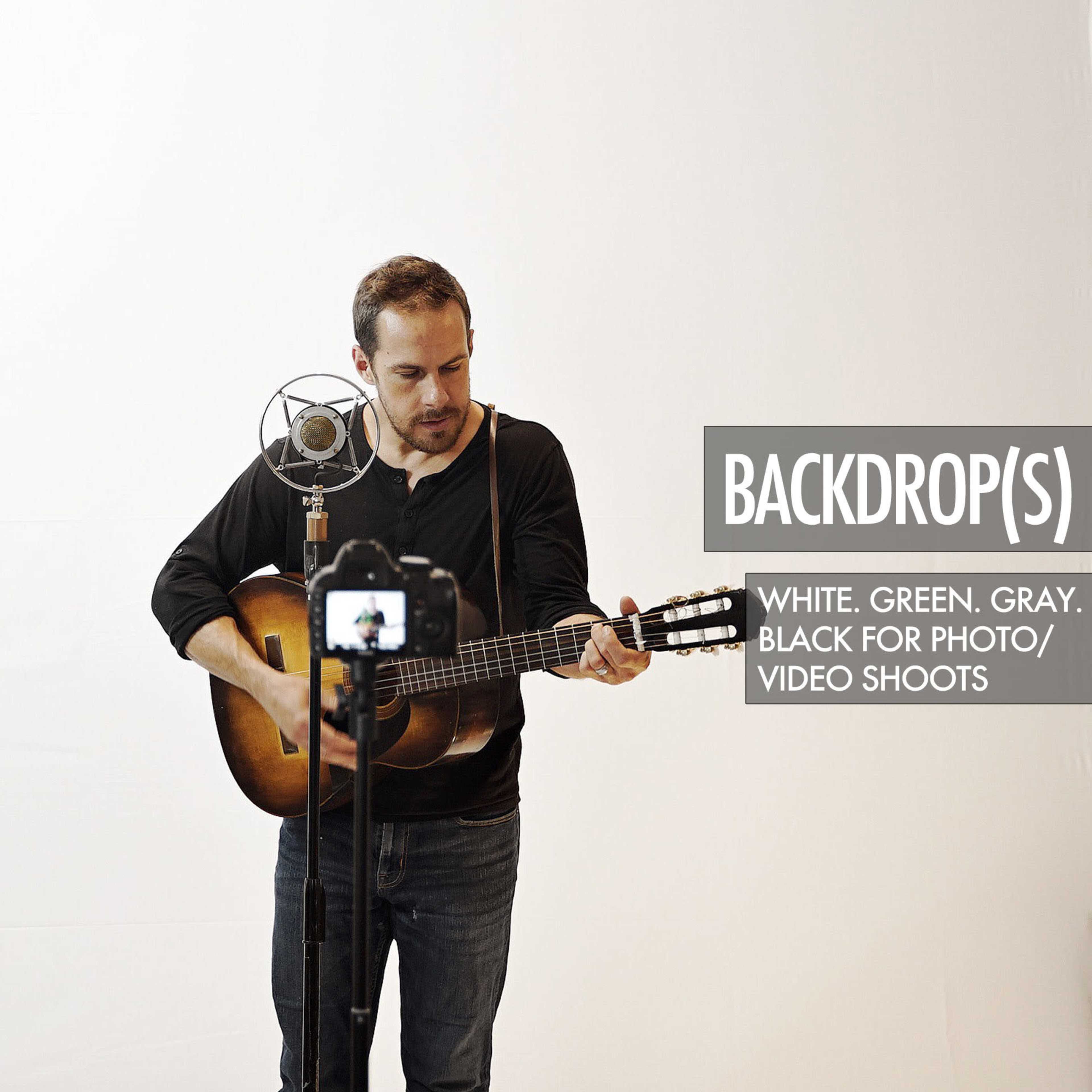 A man plays an acoustic guitar in front of a microphone against a white backdrop while another camera captures the performance.