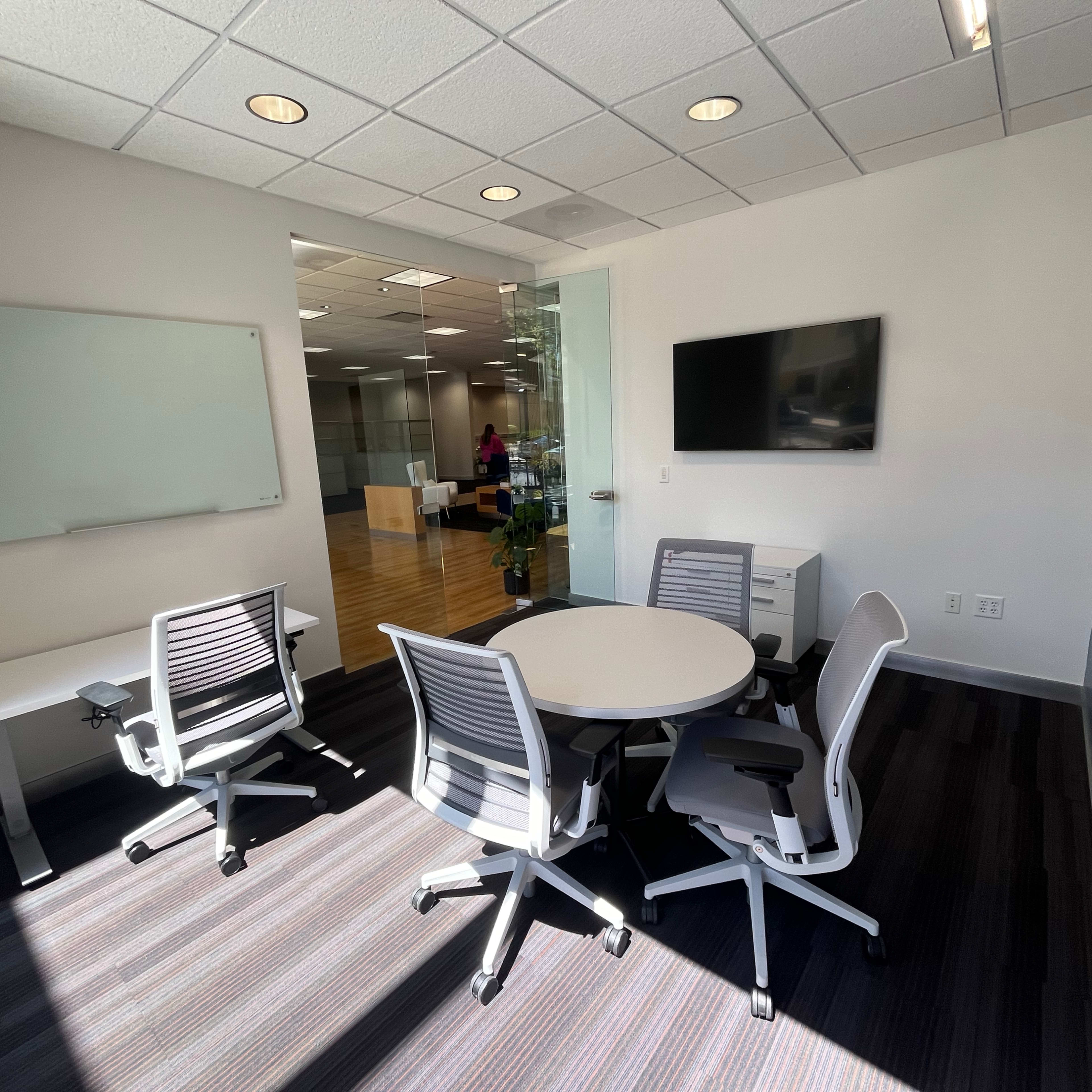 A modern office meeting room with a round table, four office chairs, a whiteboard, and a wall-mounted television.
