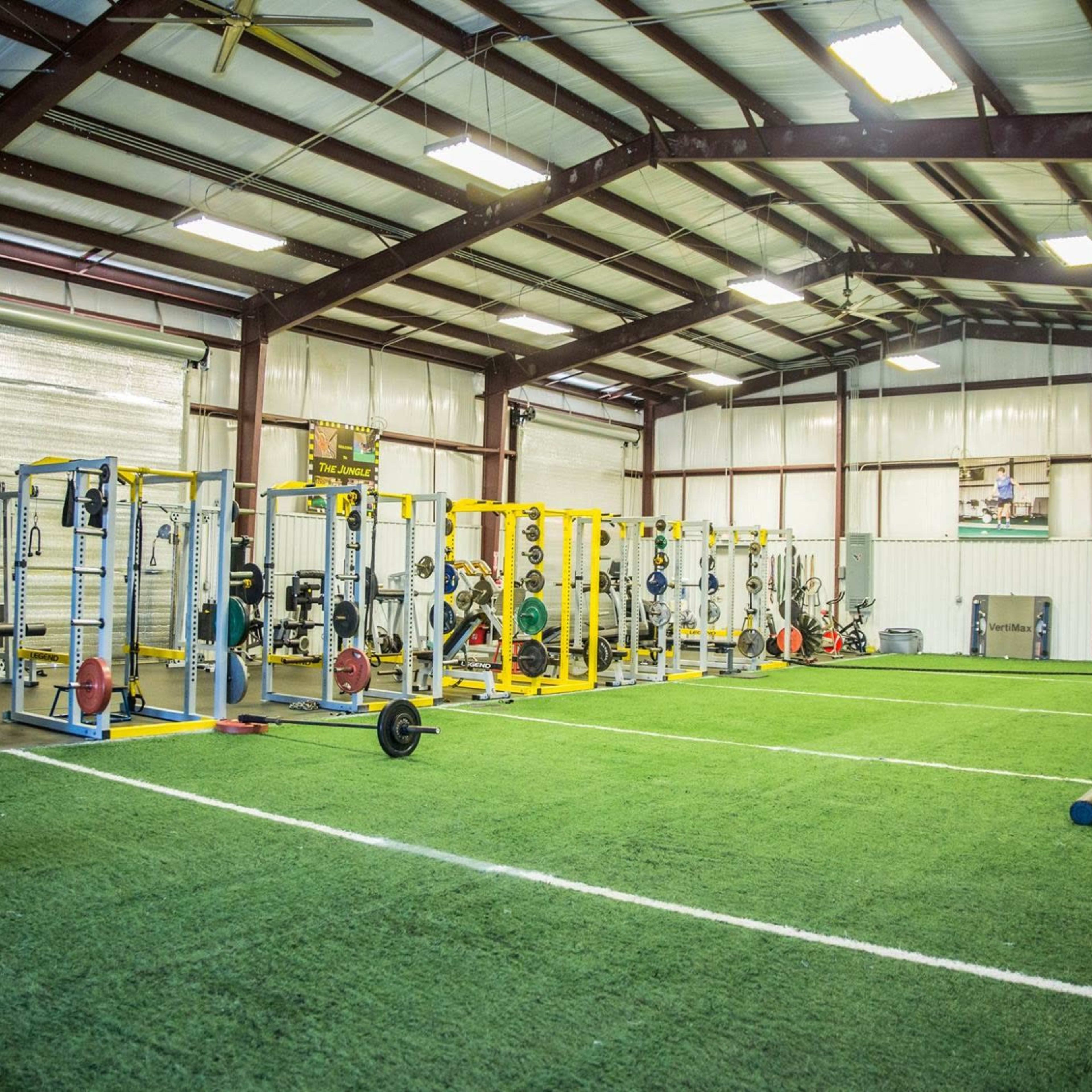 A spacious indoor gymnasium equipped with weightlifting stations, exercise equipment, and artificial turf flooring.