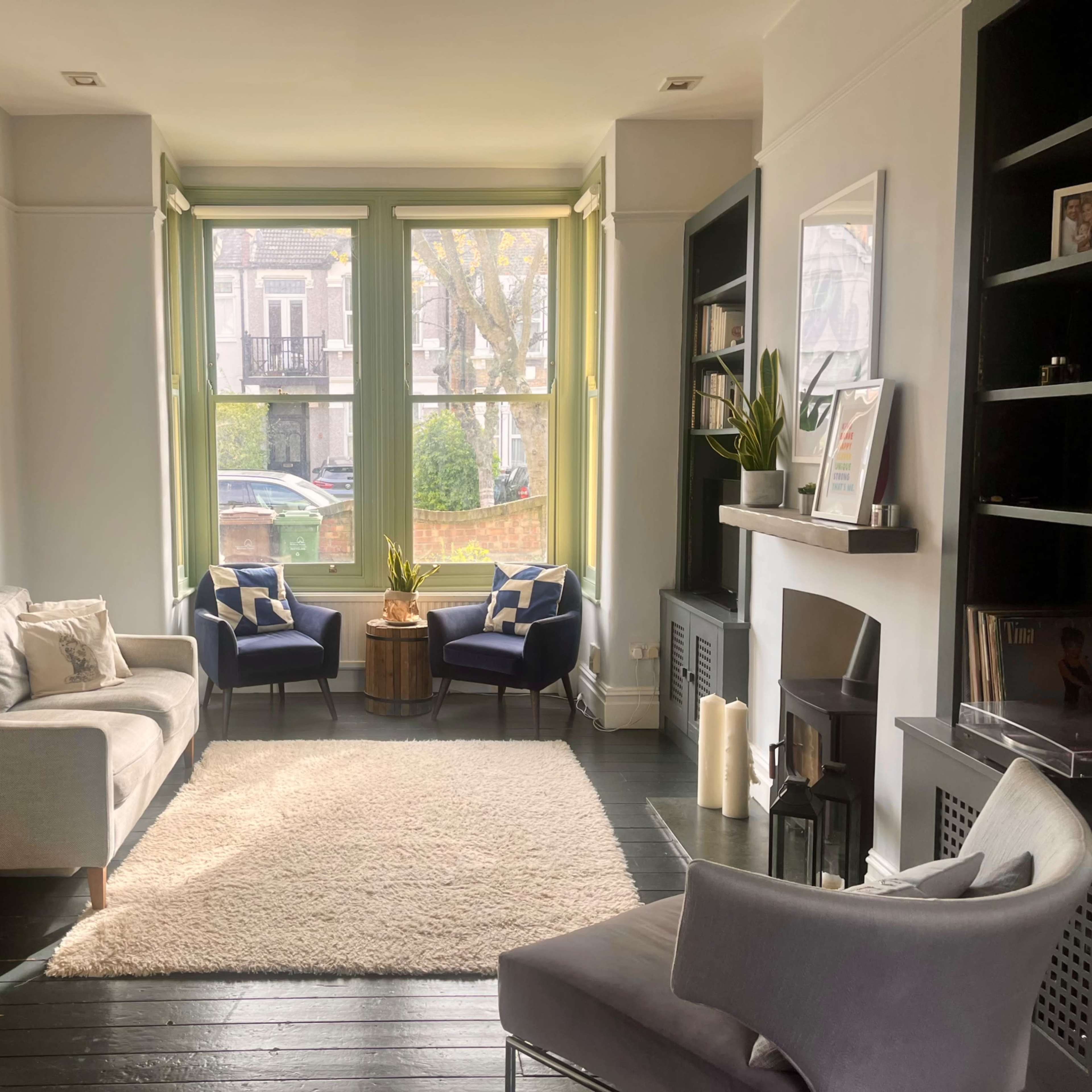 A light-filled living room features a gray sofa, two armchairs, a coffee table, and a fireplace, with large windows overlooking a quiet street.