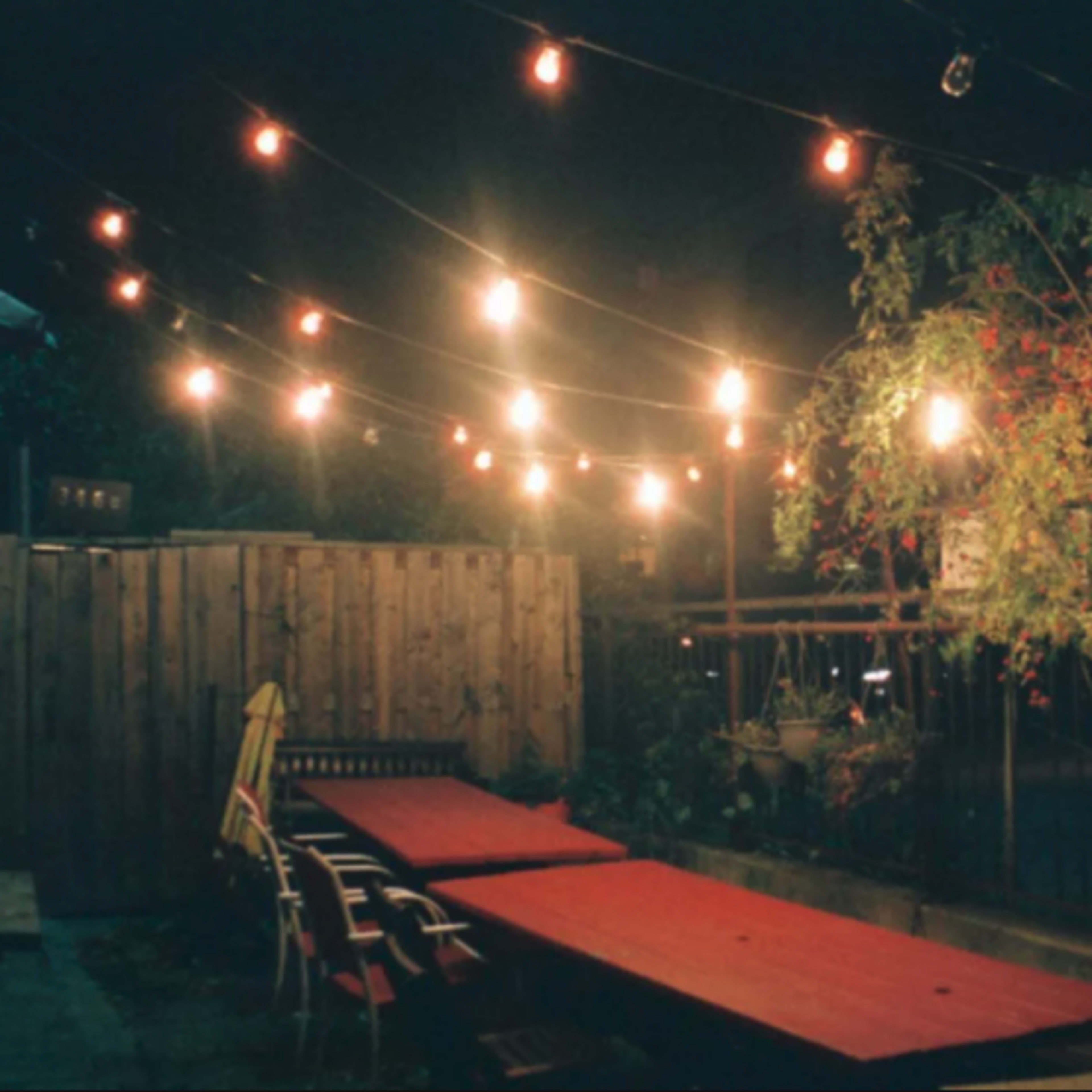 A backyard patio at night, illuminated by string lights above a long red table and wooden chairs.