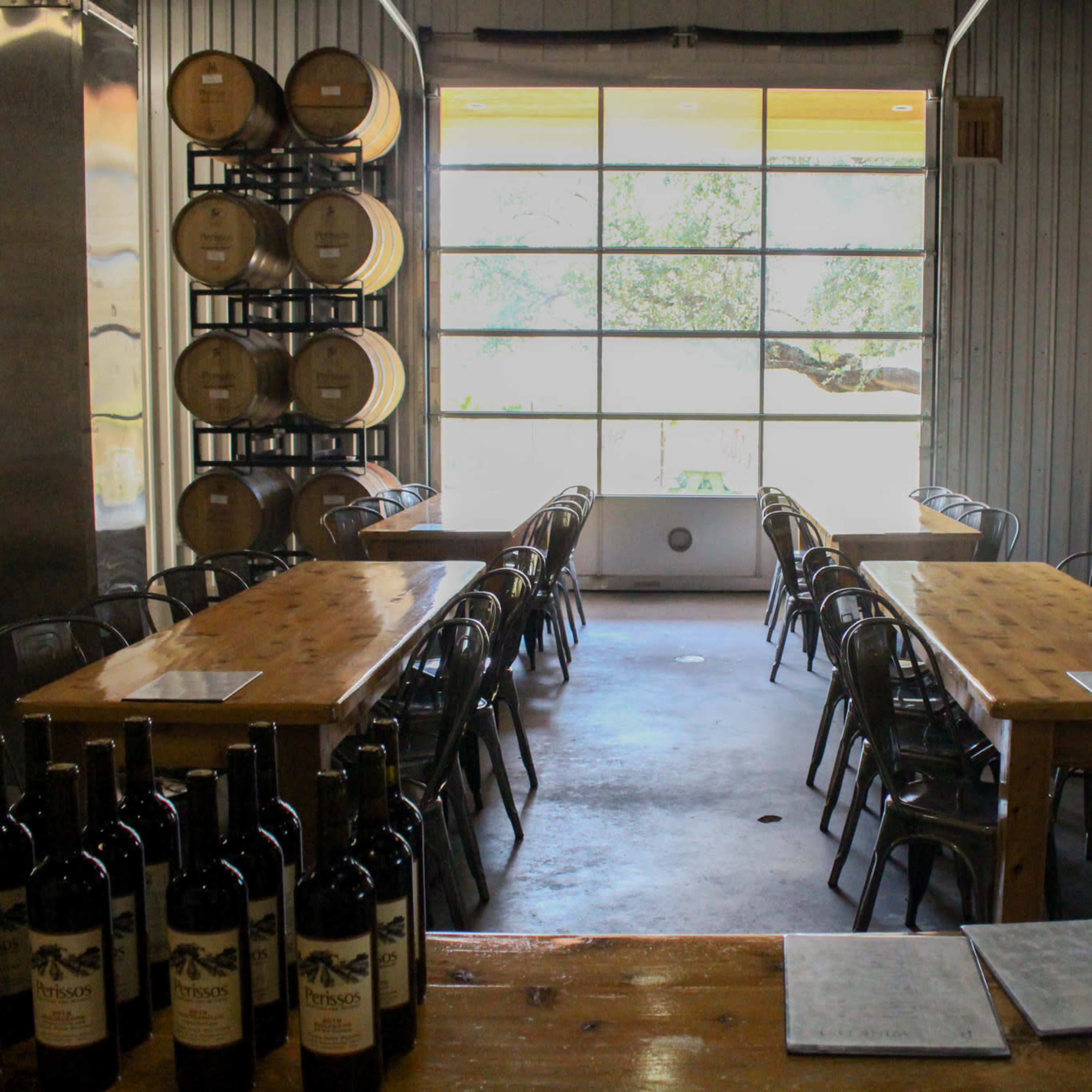 A winery tasting room with wooden tables and chairs arranged for visitors, flanked by wine barrels and large windows providing a view of the outdoors.