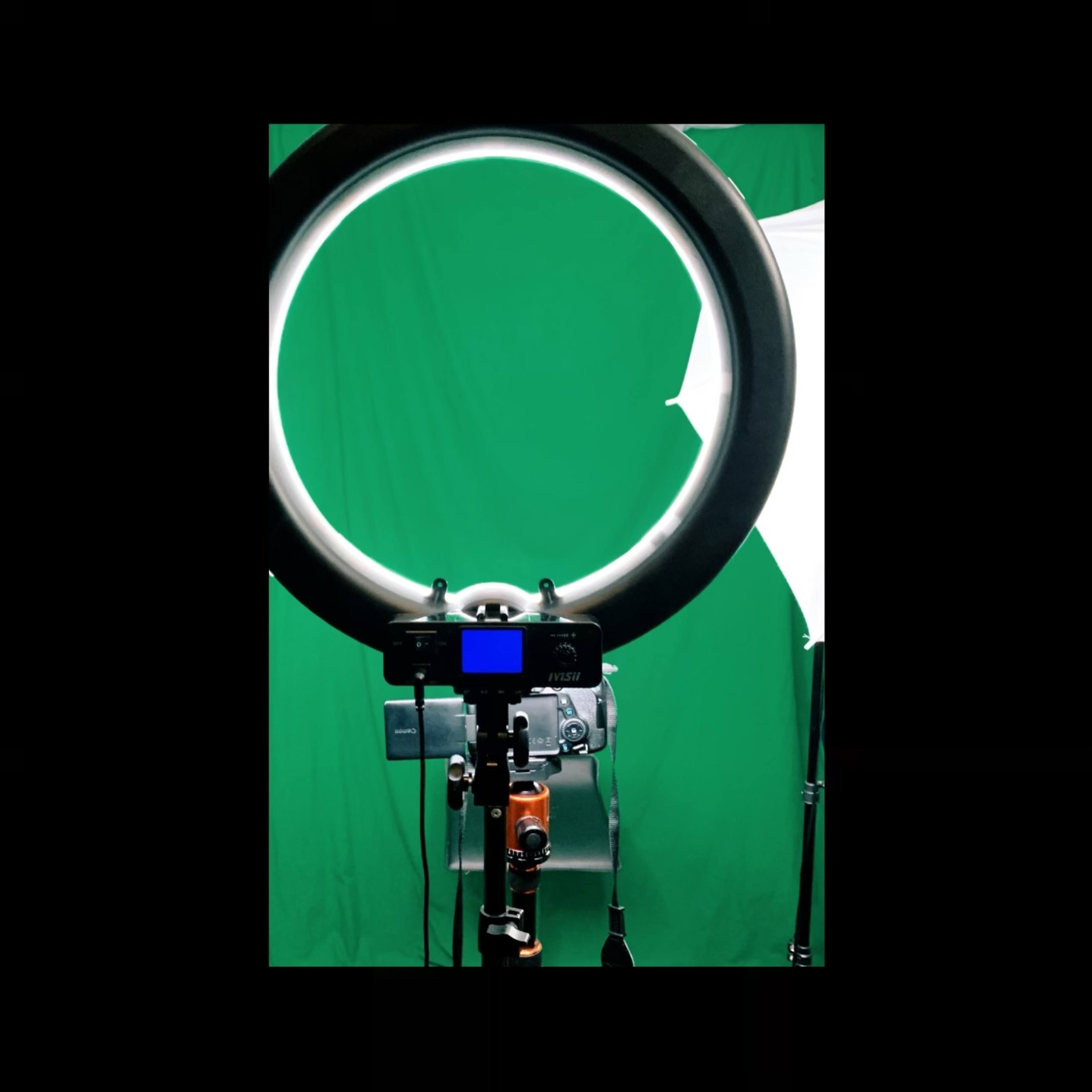 A camera is positioned in front of a ring light with a green backdrop in the background.