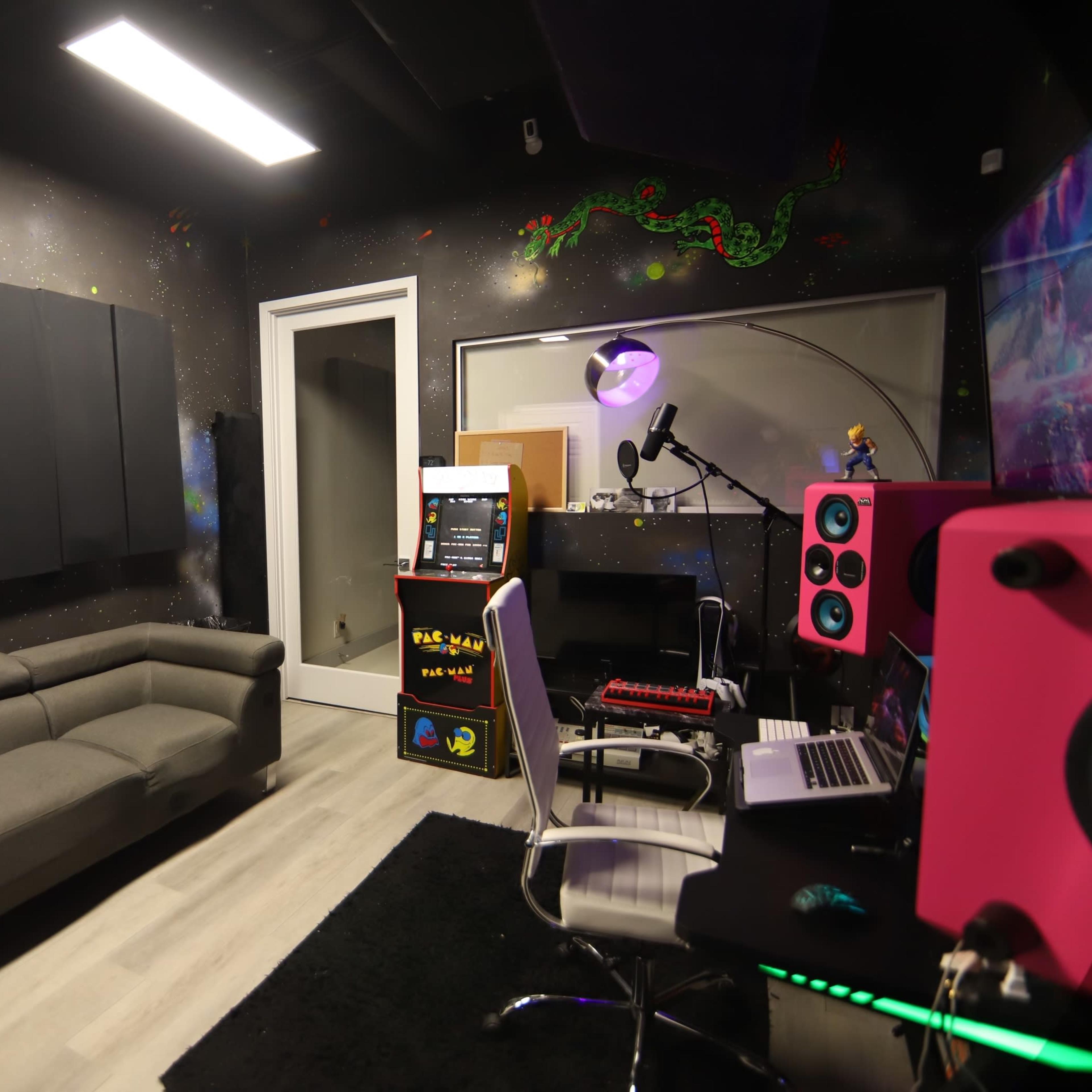 A soundproof recording studio featuring a gray sofa, a computer workstation with pink speakers, and arcade game equipment against a colorful, painted wall.