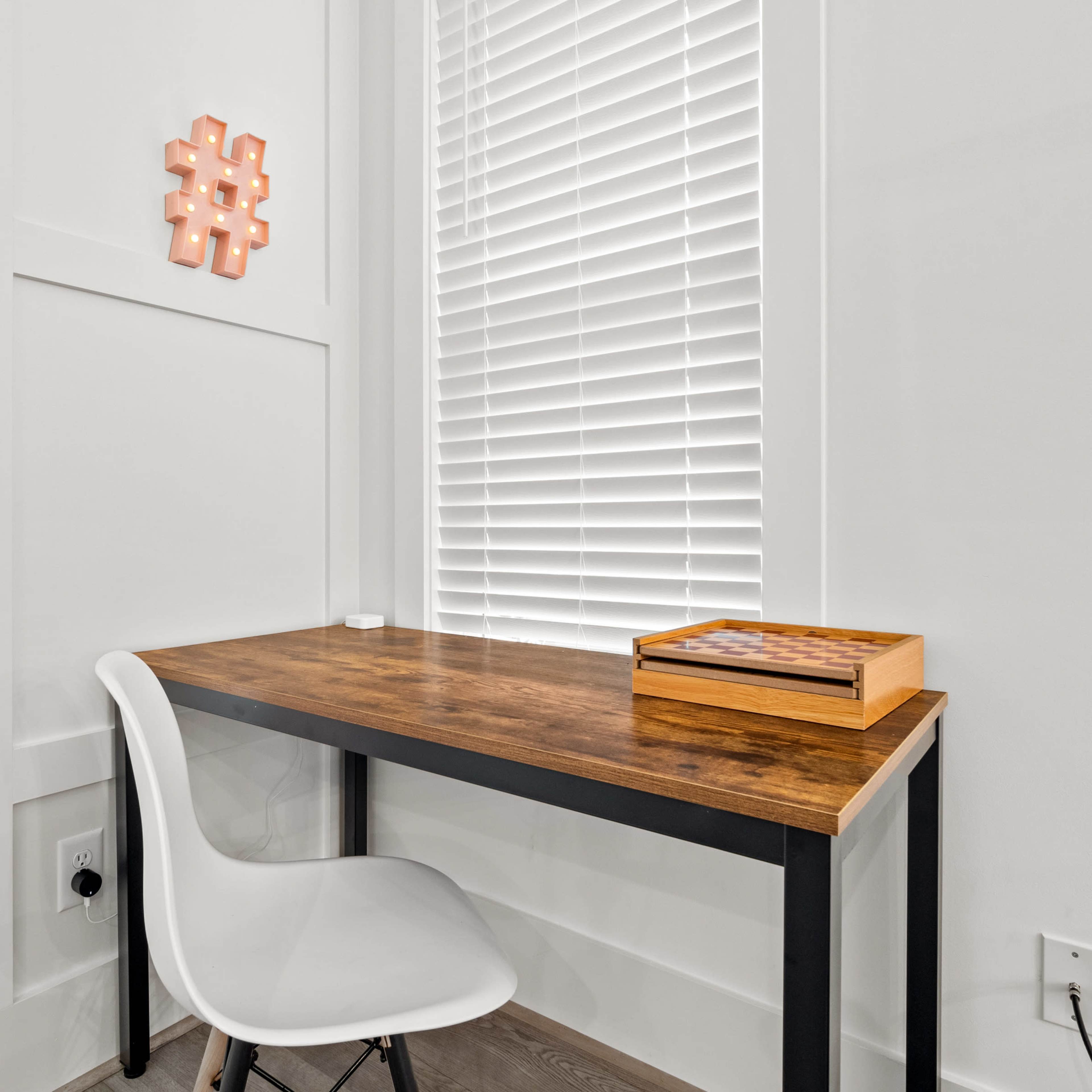 A small wooden desk with a white chair is positioned against a wall with white blinds and a decorative hashtag on display.
