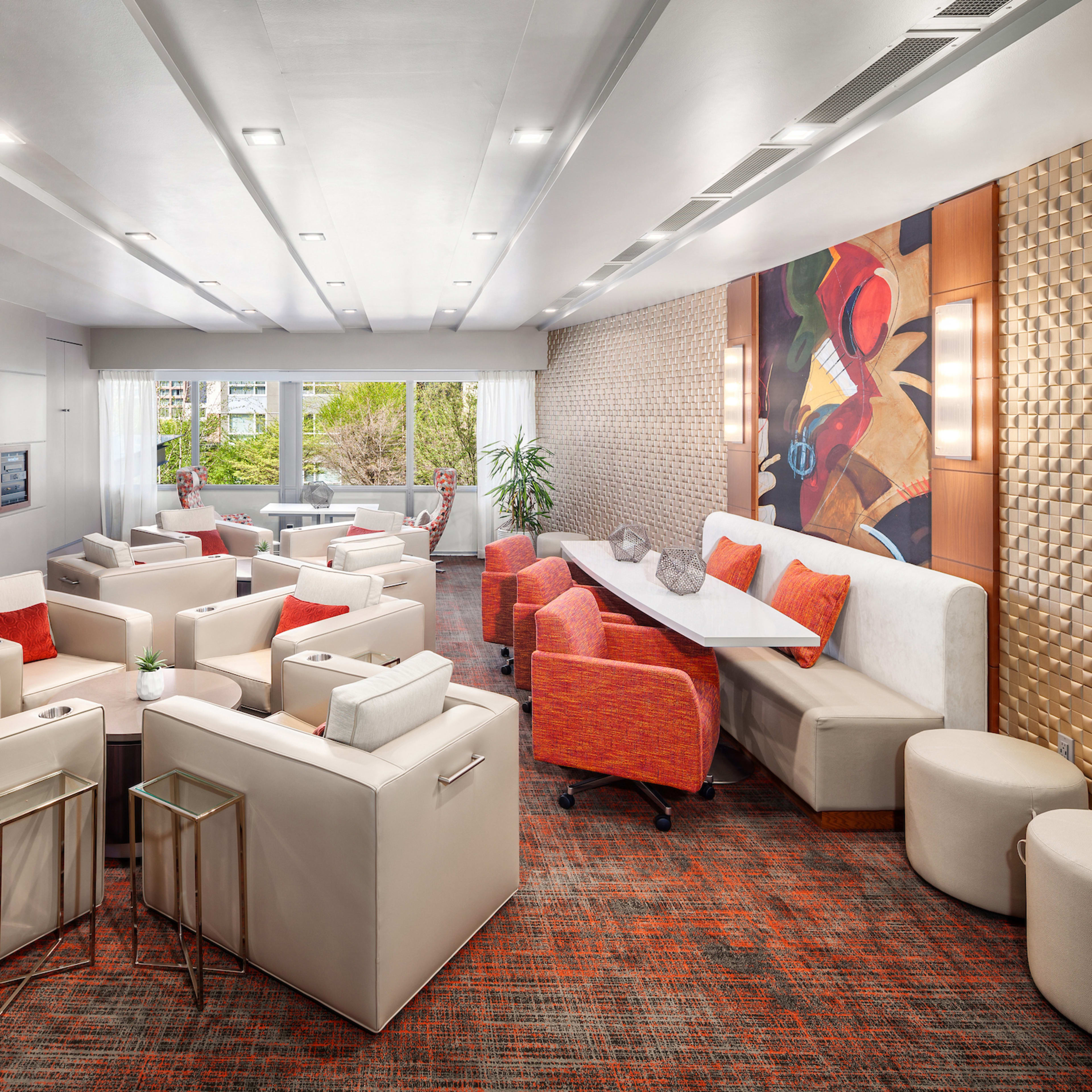 A modern, well-lit lounge area featuring comfortable seating arrangements, a large screen on the wall, and large windows providing a view of the outdoors.
