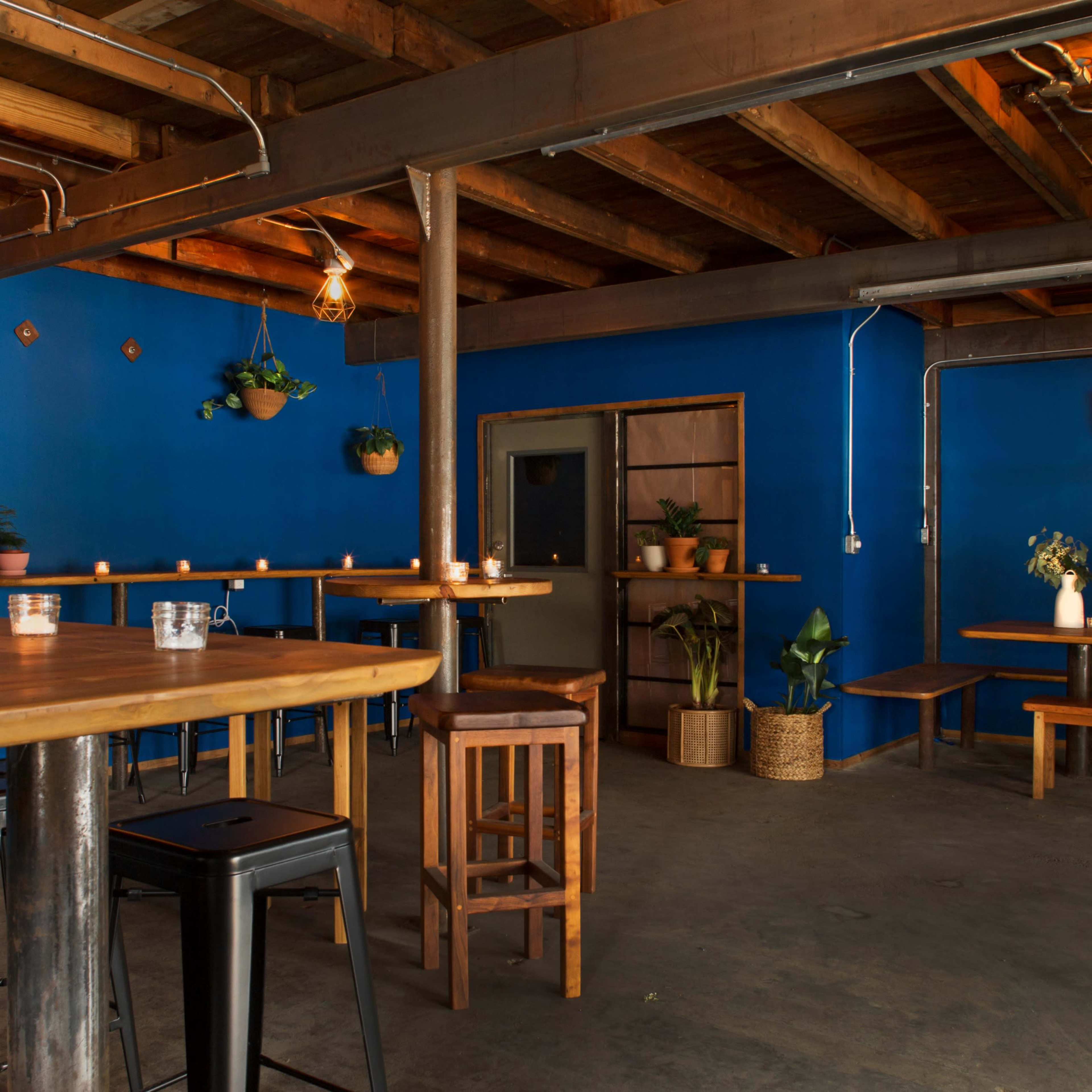 A rustic interior of a bar or café with wooden tables, benches, and a blue wall, illuminated by warm lights.
