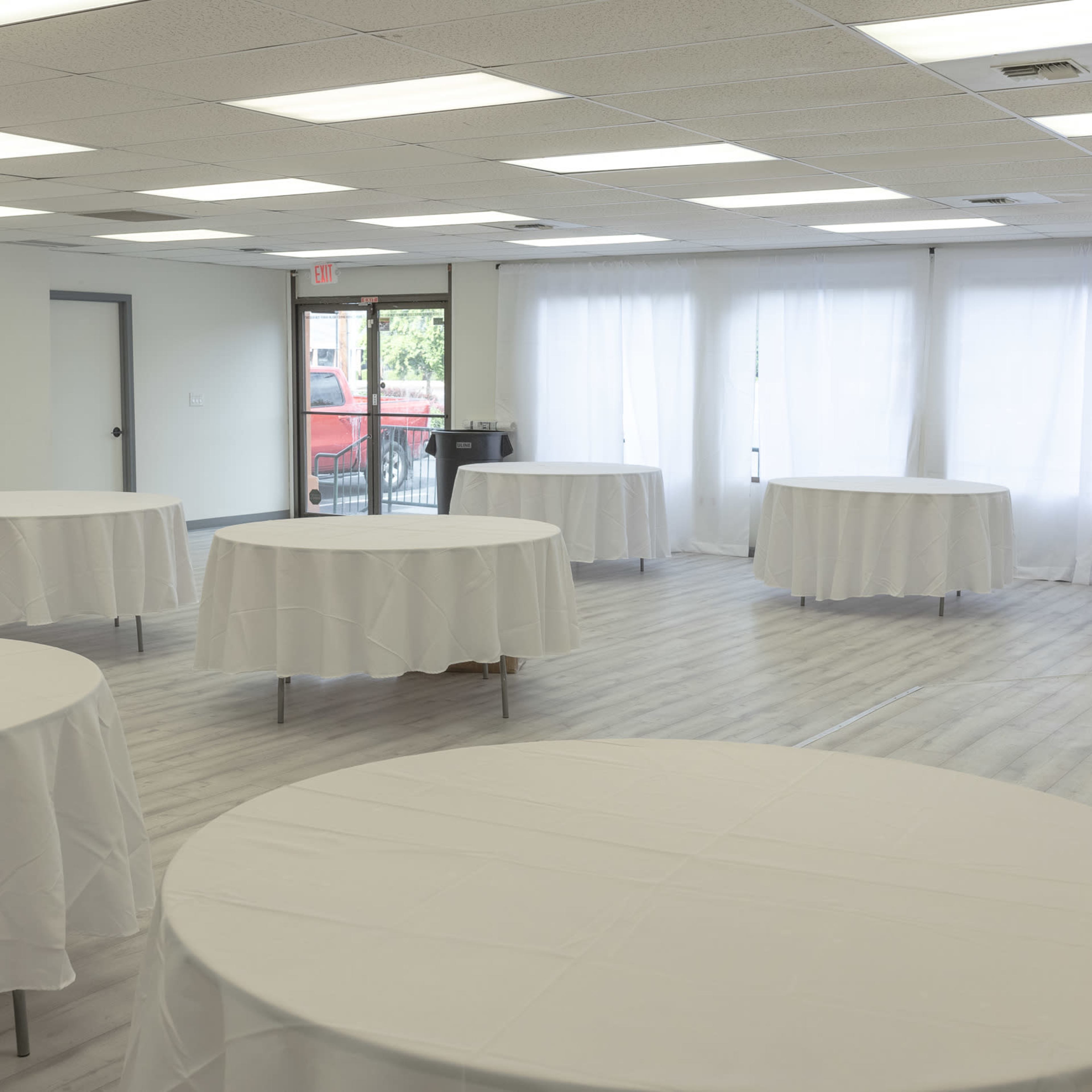 A spacious room with multiple round tables covered in white tablecloths arranged throughout the area, illuminated by overhead lighting and large windows.