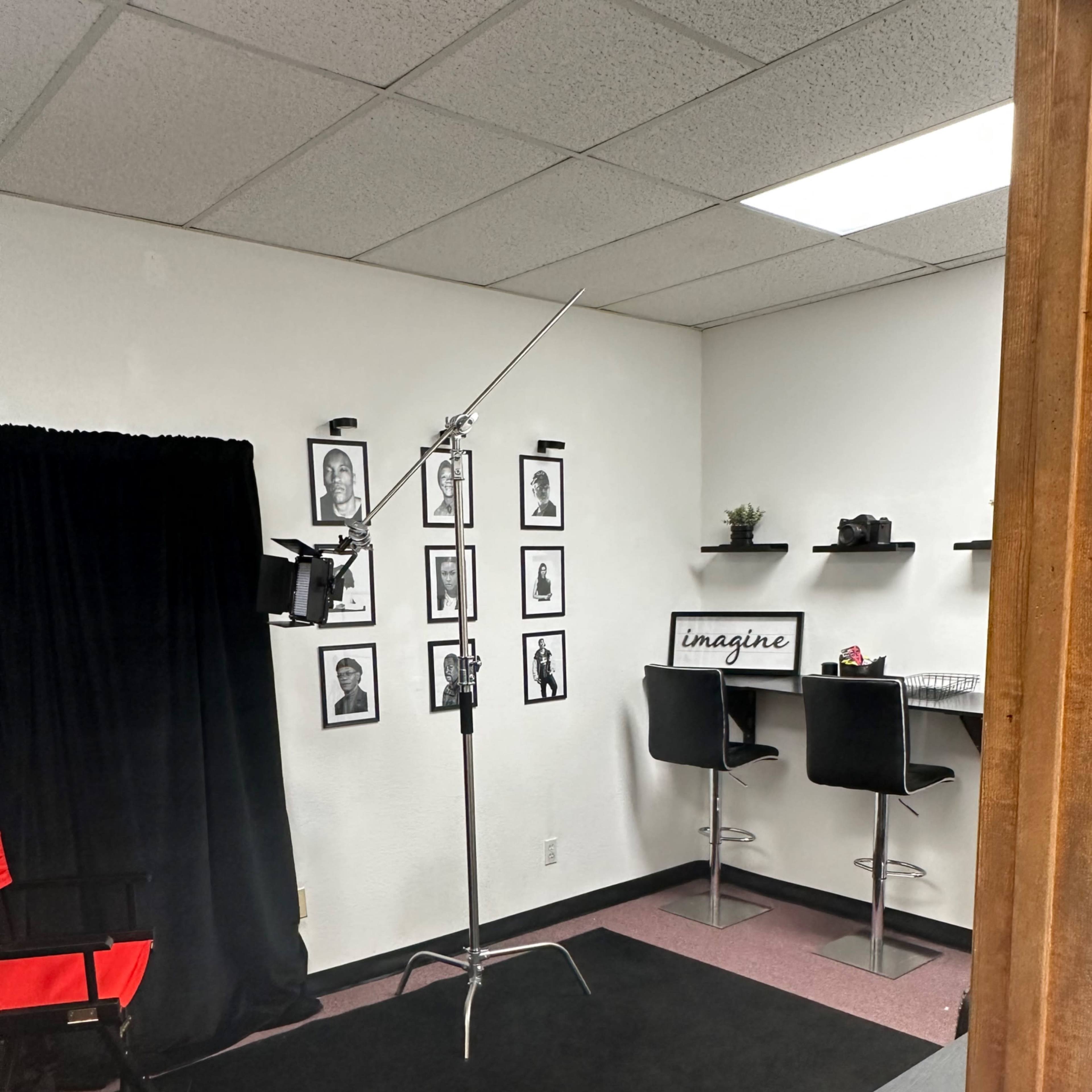 A small studio space with a black backdrop, framed photographs on the wall, a standing light on a stand, and a small counter with chairs.