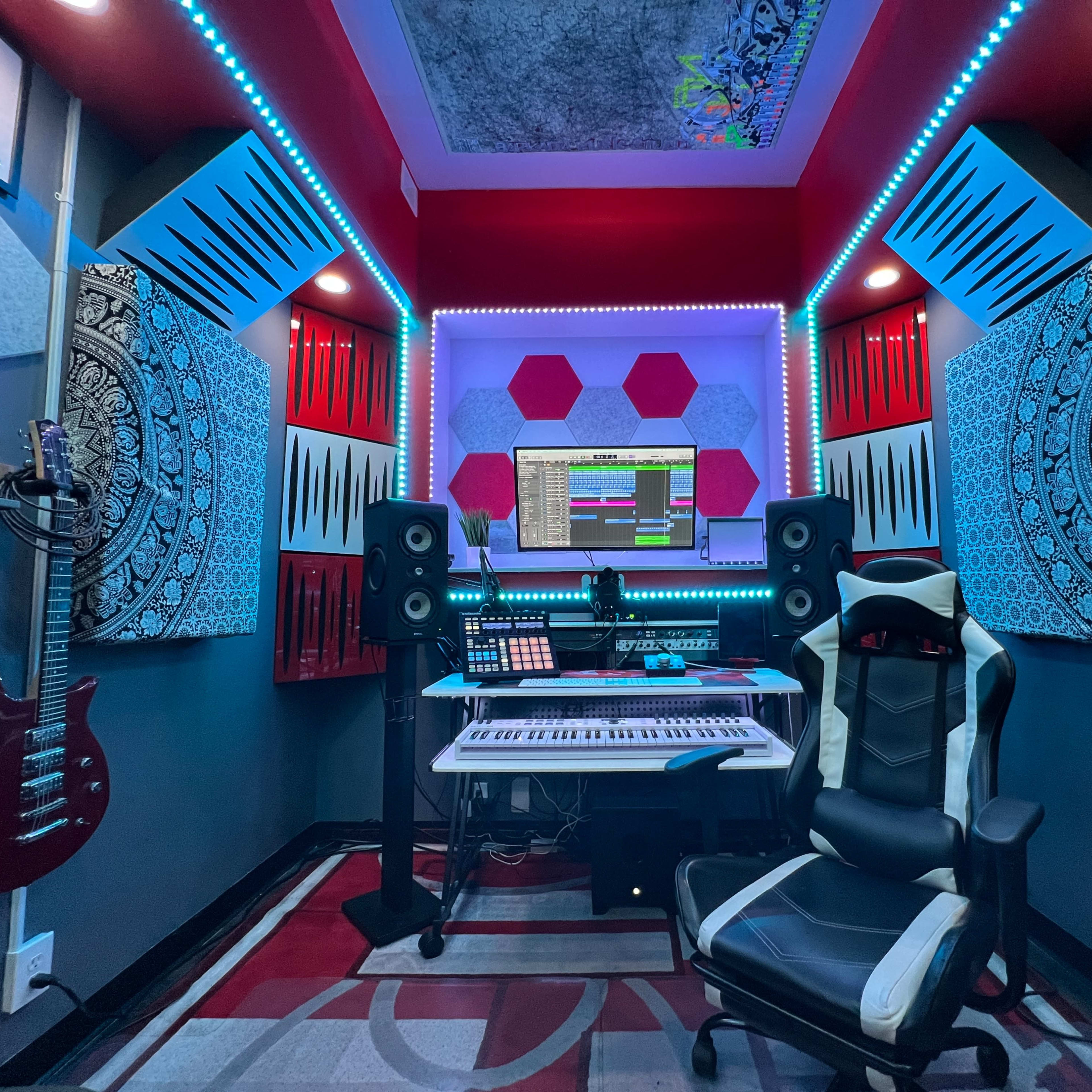 A compact music production studio featuring a keyboard, sound speakers, audio mixer, and electric guitars arranged against illuminated acoustic panels.