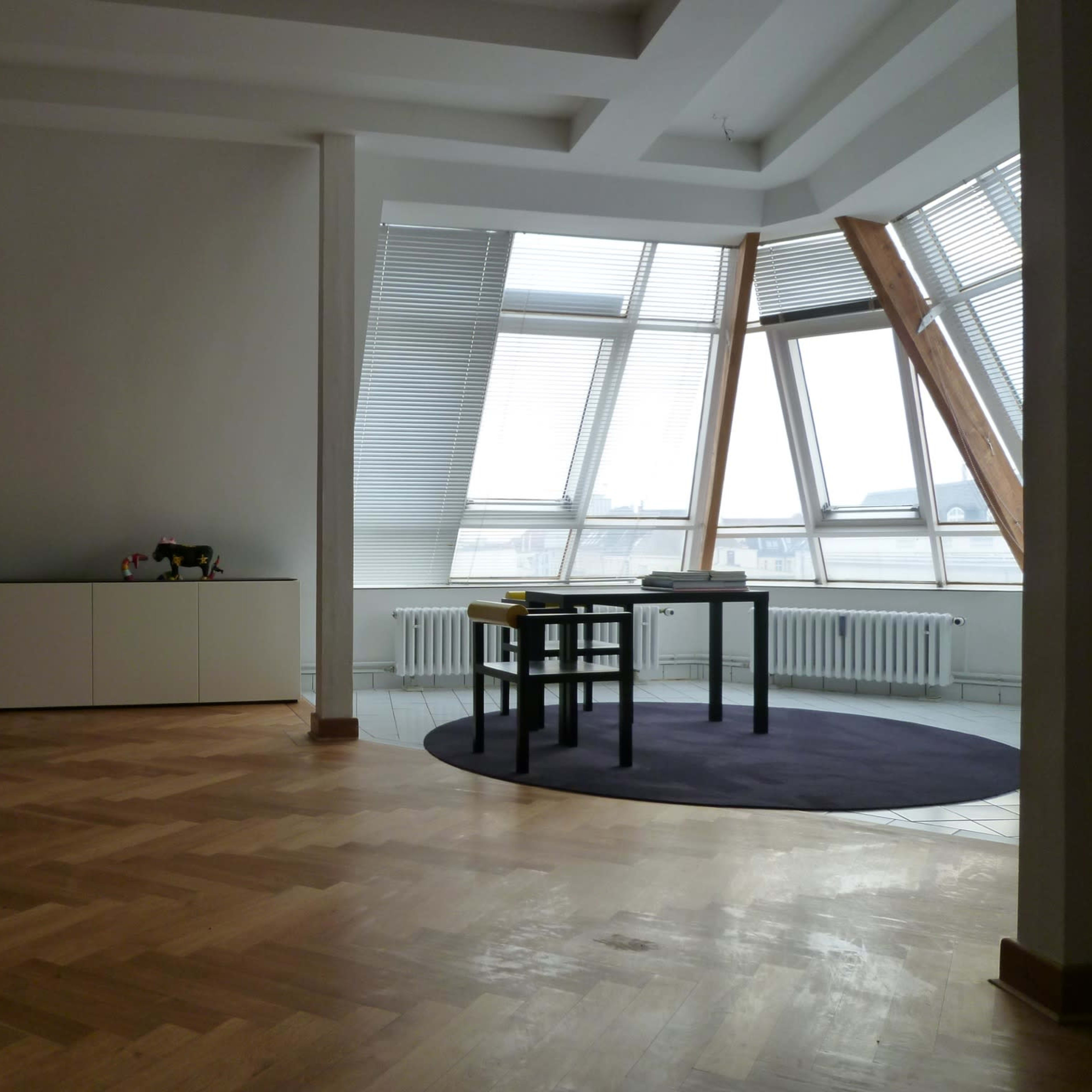 A spacious room features a table and chairs near large windows with an angular design, set on a hardwood floor.