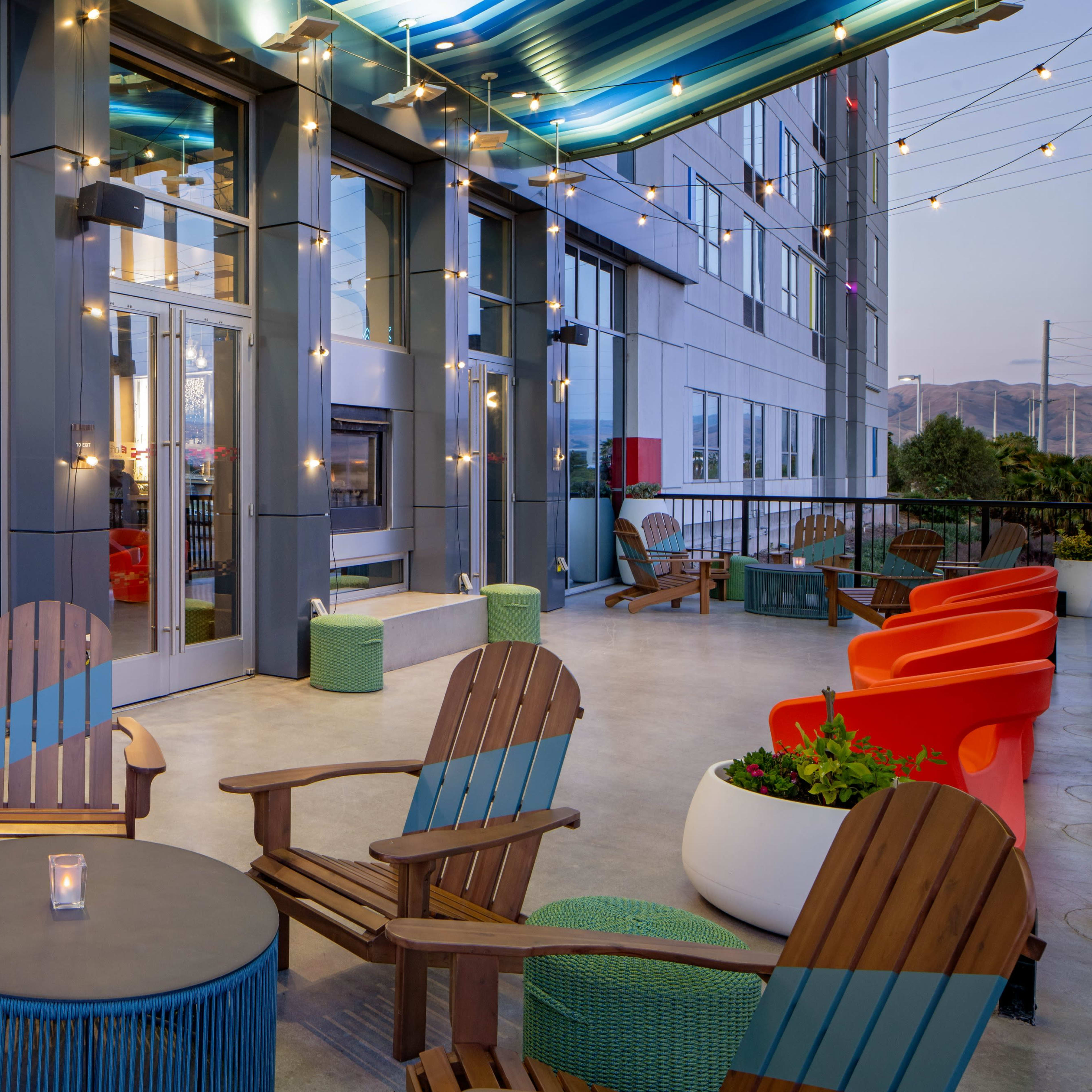 A modern outdoor lounge area with wooden chairs, colorful seating, and string lights hanging above.