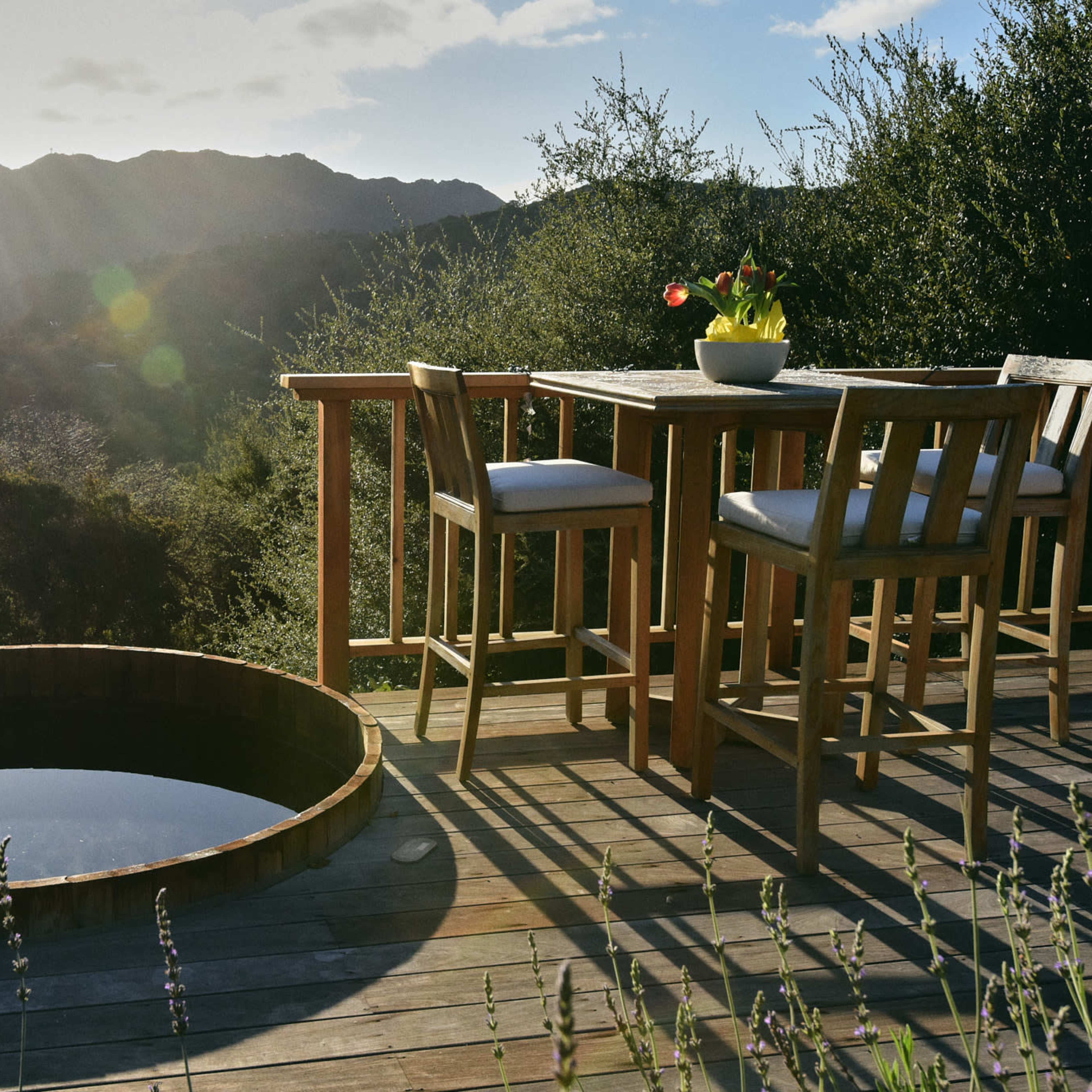 A wooden deck features a round hot tub and a dining table set against a backdrop of mountains and a setting sun.