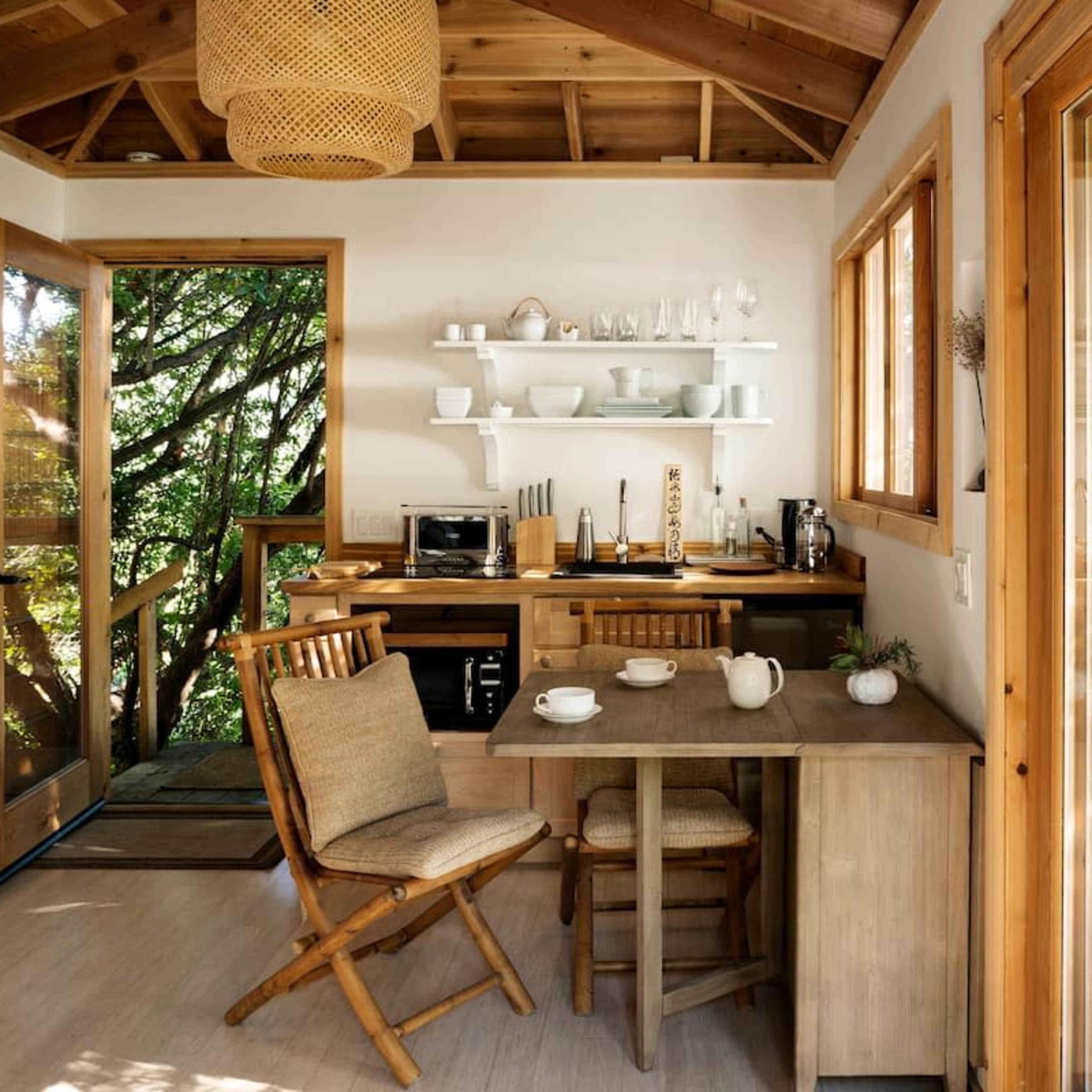 A cozy kitchen area features wooden furniture, shelves displaying dishware, and large windows overlooking a lush outdoor setting.