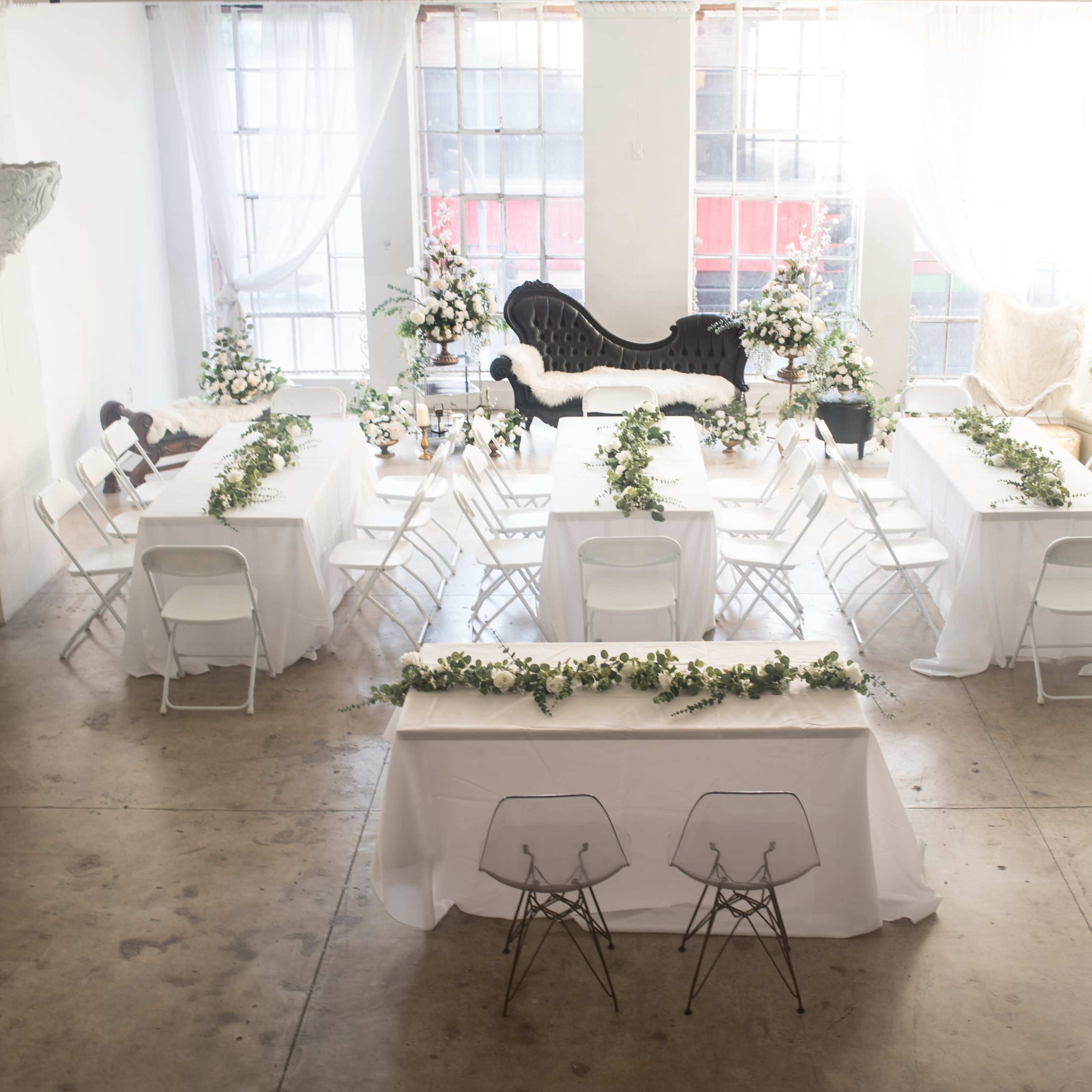A bright, spacious event room set with several white tables and chairs, adorned with greenery and floral arrangements.